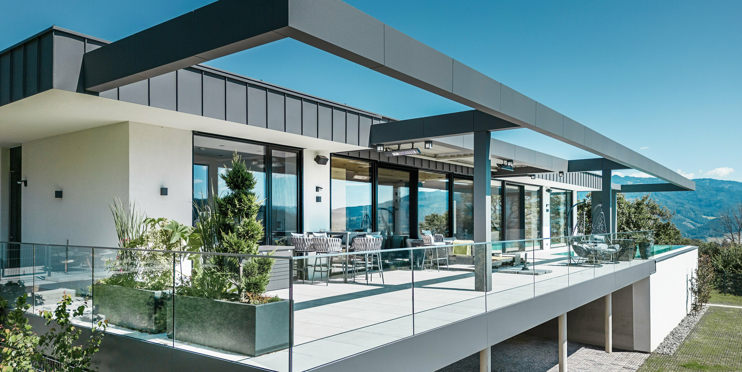 Luxusní terasa rodinného domu v rakouském Pogier s elegantním střešním a fasádním systémem od PREFA. Kombinace PREFALZ a PREFABOND v tmavě šedé barvě vytváří moderní a sofistikovaný design. Prostorná terasa nabízí dechberoucí výhled a je vybavena skleněným zábradlím, které vytváří otevřenou a příjemnou atmosféru. Částečně zastřešený venkovní prostor umožňuje vychutnat si krásy okolní přírody a je ideální pro relaxaci a společenská setkání.