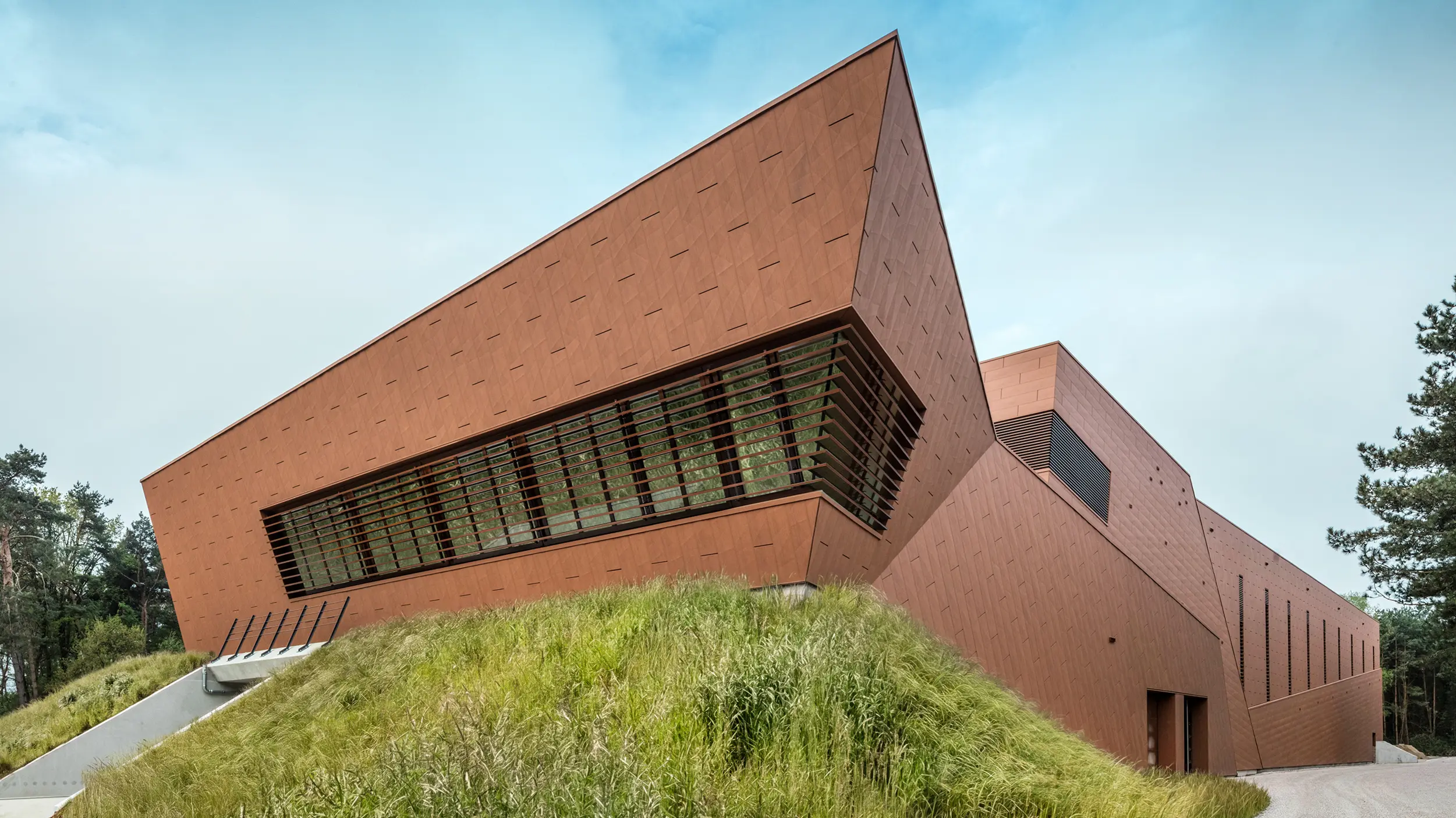 La remarquable station de pompage d'eau potable de WML à Lottum se distingue par sa forme sculpturale inclinée et sa façade en aluminium PREFA Siding.X, d'une teinte rouille chaleureuse. La surface à la structure dynamique reflète la lumière et crée de forts accents architecturaux dans le paysage. De larges ouvertures à persiennes et des lignes épurées intègrent harmonieusement technologie et design. Durables, légers et résistants à la corrosion, les panneaux en aluminium PREFA offrent une recyclabilité durable, des performances élevées et une enveloppe de bâtiment nécessitant peu d'entretien – un atout architectural moderne pour les infrastructures d'avenir.