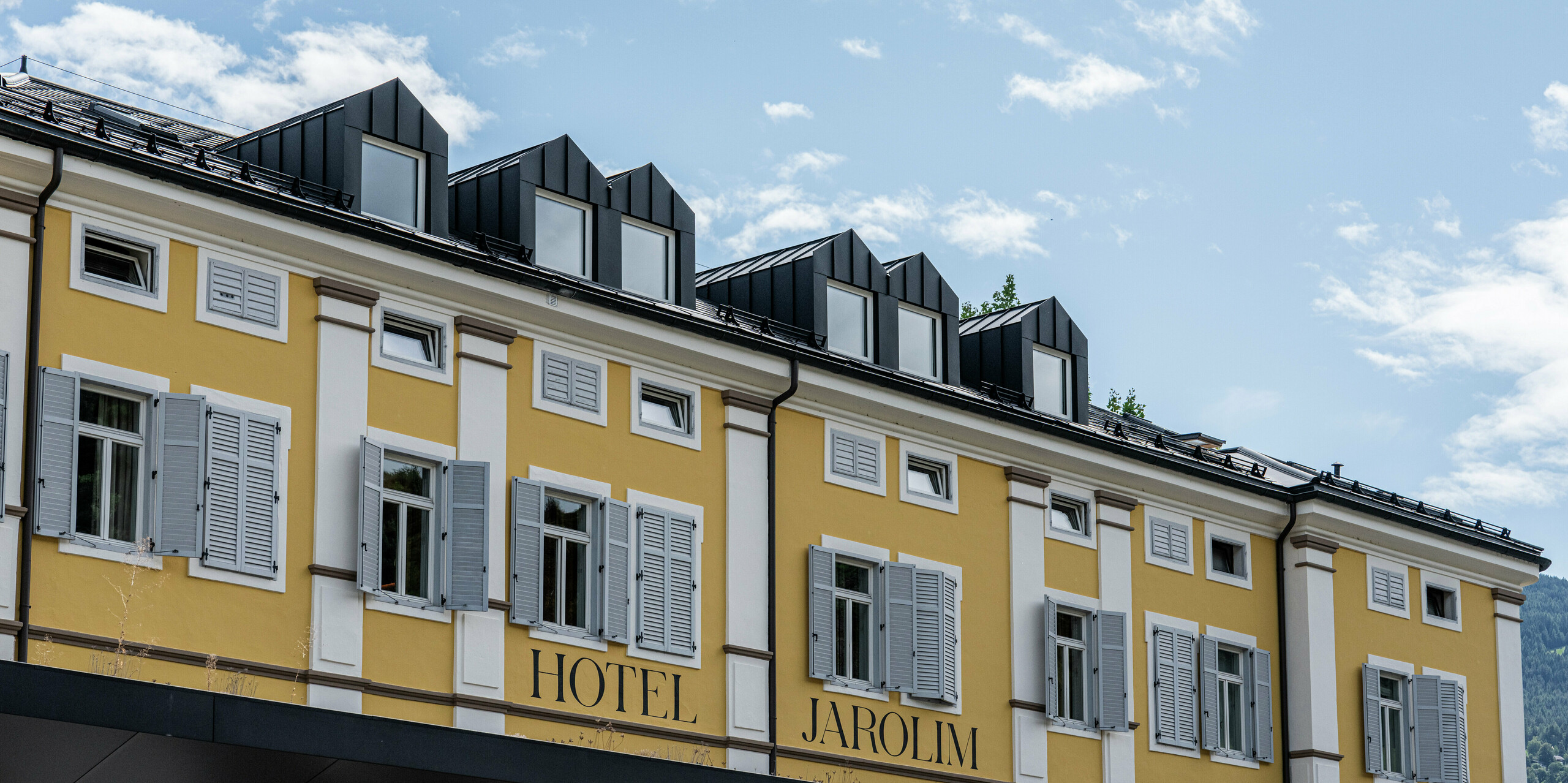 Vue détaillée de l'hôtel Jarolim à Brixen avec des lucarnes modernes revêtues d'aluminium PREFA. Les lucarnes fabriquées avec précision et les R.16 en P.10 noir soulignent l'élégance et la fonctionnalité du toit. Le toit PREFA, durable et résistant aux intempéries, s'intègre parfaitement à l'architecture historique et allie tradition et innovation. Le choix parfait pour des solutions de toiture durables, esthétiques et de haute qualité.