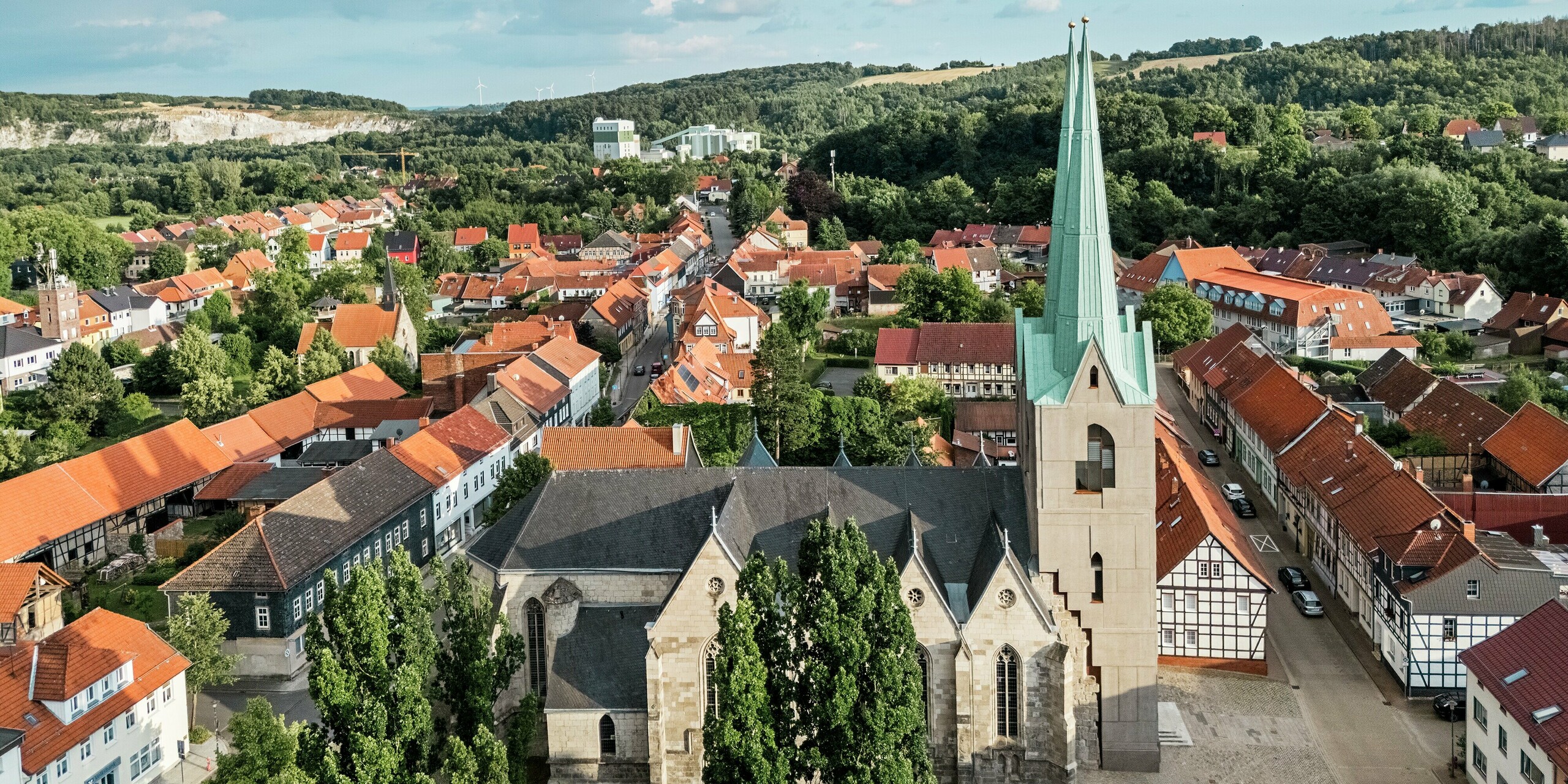 Drohnenaufnahme der Stadtkirche St. Johannis in Ellrich, eingebettet in das malerische Stadtbild mit roten Ziegeldächern. Im Zentrum: der neu errichtete Turm mit spitz zulaufendem Dach aus PREFALZ in P.10 Patinagrün – ein moderner Eyecatcher mit historischem Bezug. Das hochwertige Aluminiumdach überzeugt durch Langlebigkeit, Sturmfestigkeit und wartungsarme Eleganz.