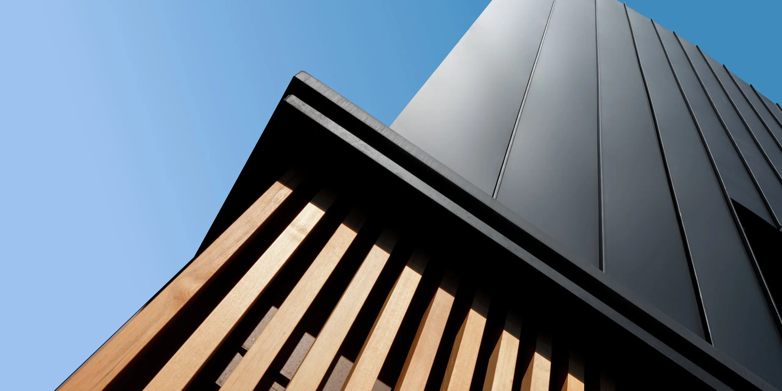 Détail architectural d'une maison jumelée moderne à Cattolica : un bardage vertical en aluminium PREFALZ gris noir s'harmonise avec d'élégantes lames de bois. La pureté des lignes et la qualité de la surface PREFA garantissent durabilité, protection contre la corrosion et un design saisissant sous un ciel d'azur.
