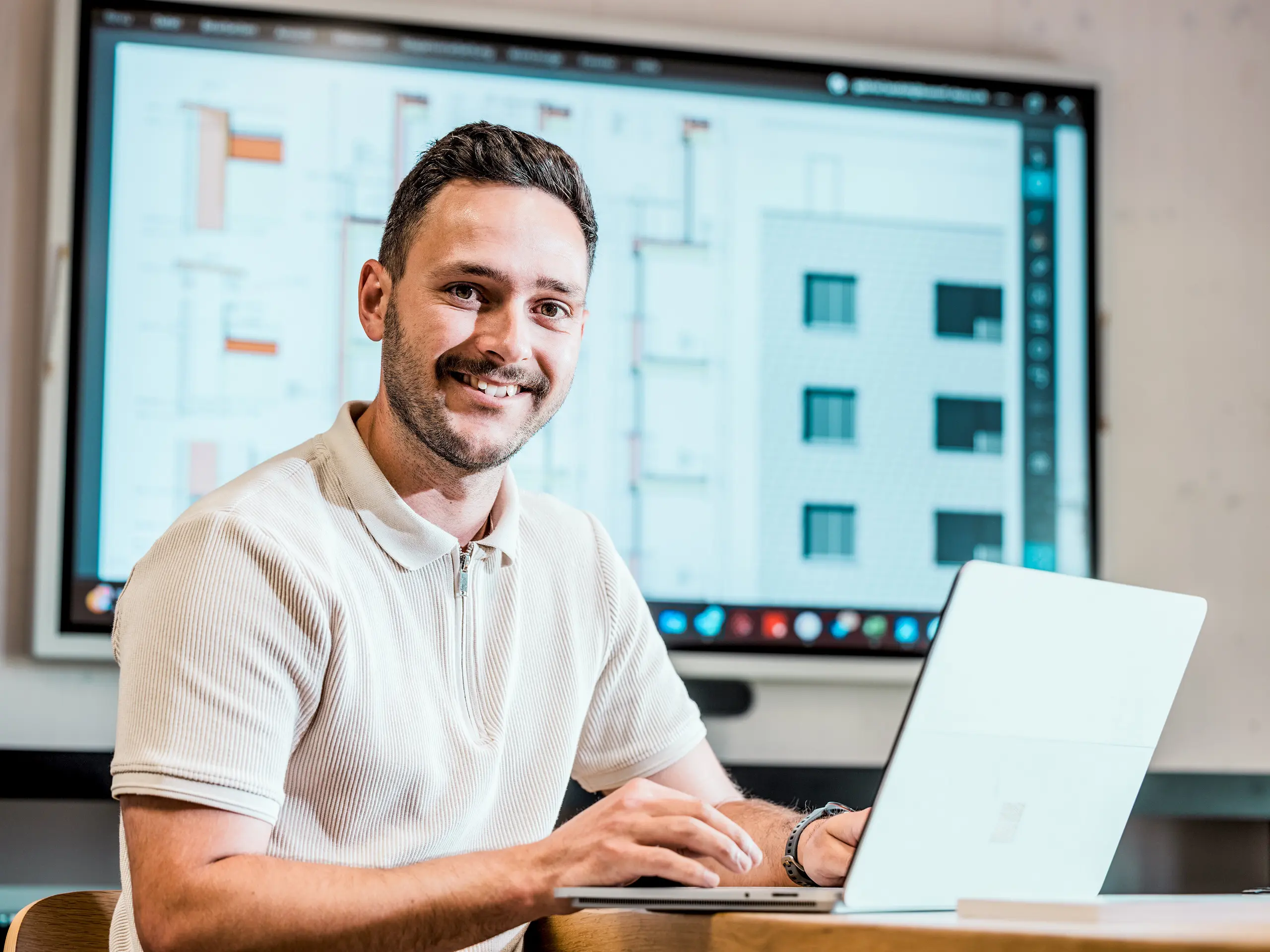 Portrait de Gabriel Landolt, employé de Künzli Davos AG, devant son ordinateur portable et un grand écran affichant les plans numériques d'un bâtiment et les détails de sa façade. L'écran présente les dessins techniques et les élévations d'un bâtiment moderne à la façade en écailles, construite avec des bardeaux PREFA couleur Gris souris P.10. Cette façade innovante en aluminium séduit par sa légèreté, sa résistance aux intempéries et sa durabilité. Une planification précise et l'utilisation de produits PREFA de haute qualité garantissent une enveloppe de bâtiment stable, nécessitant peu d'entretien et d'une grande valeur architecturale.