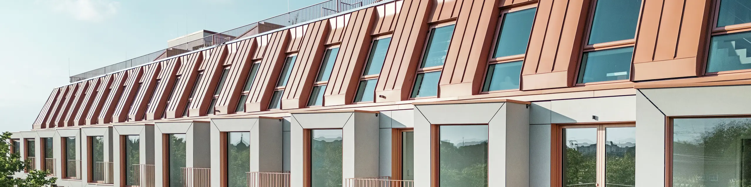 Blick auf die Fassade des Wohnprojekts Van B in München mit dem markanten PREFALZ-Aluminiumdach in der Sonderfarbe IGP Orange HDP von PREFA. Die moderne Dachkonstruktion vereint ästhetische Eleganz mit technischer Präzision und bietet dauerhaften Schutz vor Witterungseinflüssen. Kubische Erker mit großen Fensterflächen und sanft geschwungene Balkone schaffen ein harmonisches Zusammenspiel von Licht, Raum und Funktionalität – ideal für anspruchsvolle Wohnkonzepte im urbanen Raum.