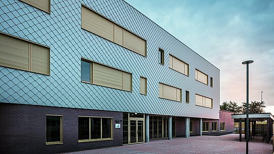 Das Foto zeigt die Eingangsseite des Collège Léontine Dolivet mit moderner Aluminiumfassade aus PREFA Wandrauten in Silbermetallic. Mehrere rechteckige Fenster mit gelben Rahmen und geschlossenen Rollläden strukturieren die Ansicht. Im Vordergrund liegt ein gepflasterter Vorplatz, flankiert von einem überdachten Bereich. Die Fassade wirkt elegant, wetterbeständig und langlebig.