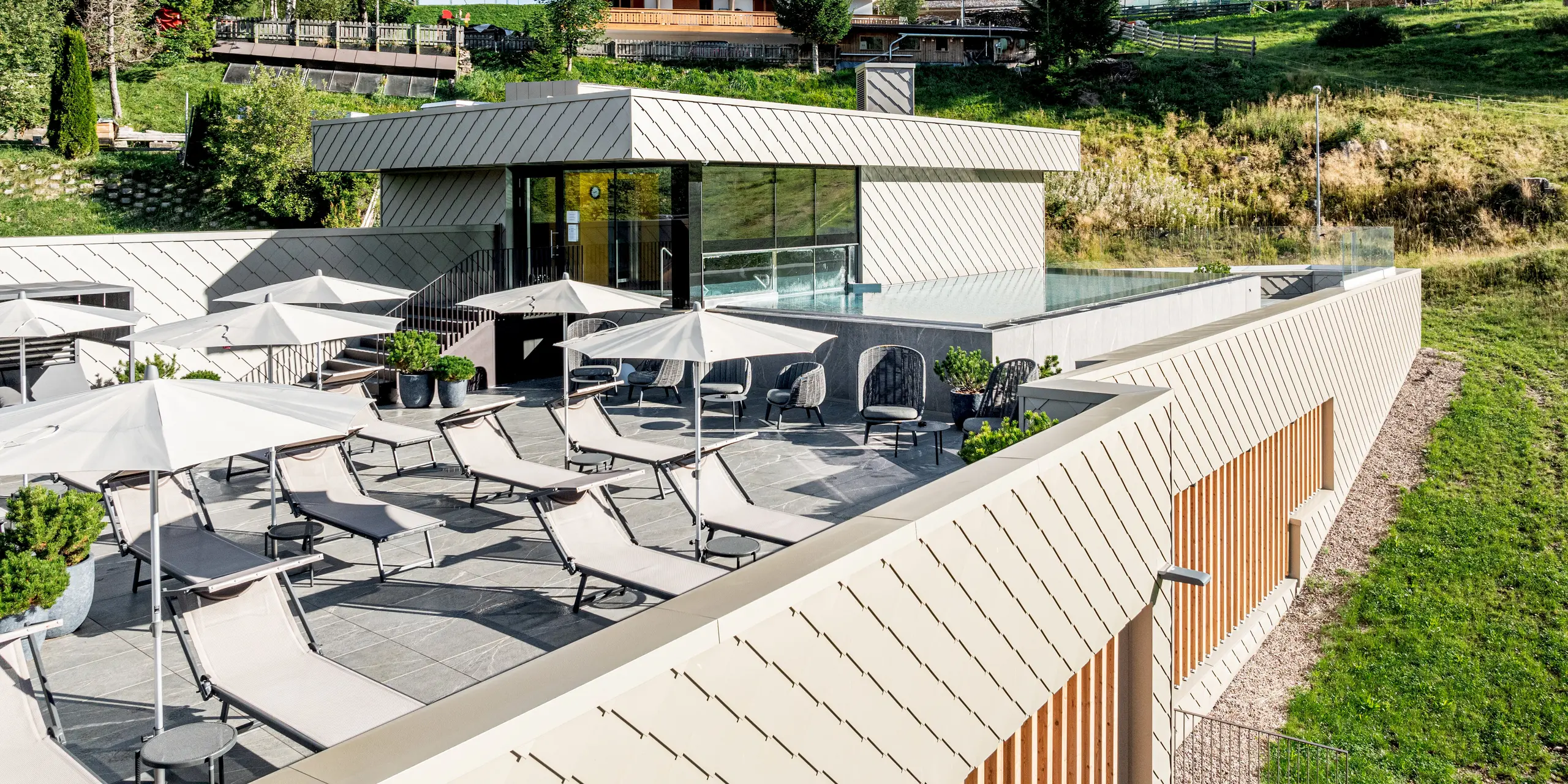 Terrasse du Sporthotel Obereggen, au Tyrol du Sud, avec piscine extérieure, transats et parasols, nichée dans un paysage alpin. L'architecture moderne est caractérisée par une façade en aluminium PREFA composée de losanges de façade 29x29 couleur bronze, aux lignes épurées et à la texture vibrante. La teinte chaude du bronze s'harmonise avec le bois et la nature. Durables, résistants aux intempéries et faciles d'entretien, les produits en aluminium PREFA offrent une grande liberté de conception et sont parfaitement adaptés à l'architecture hôtelière haut de gamme en région alpine.