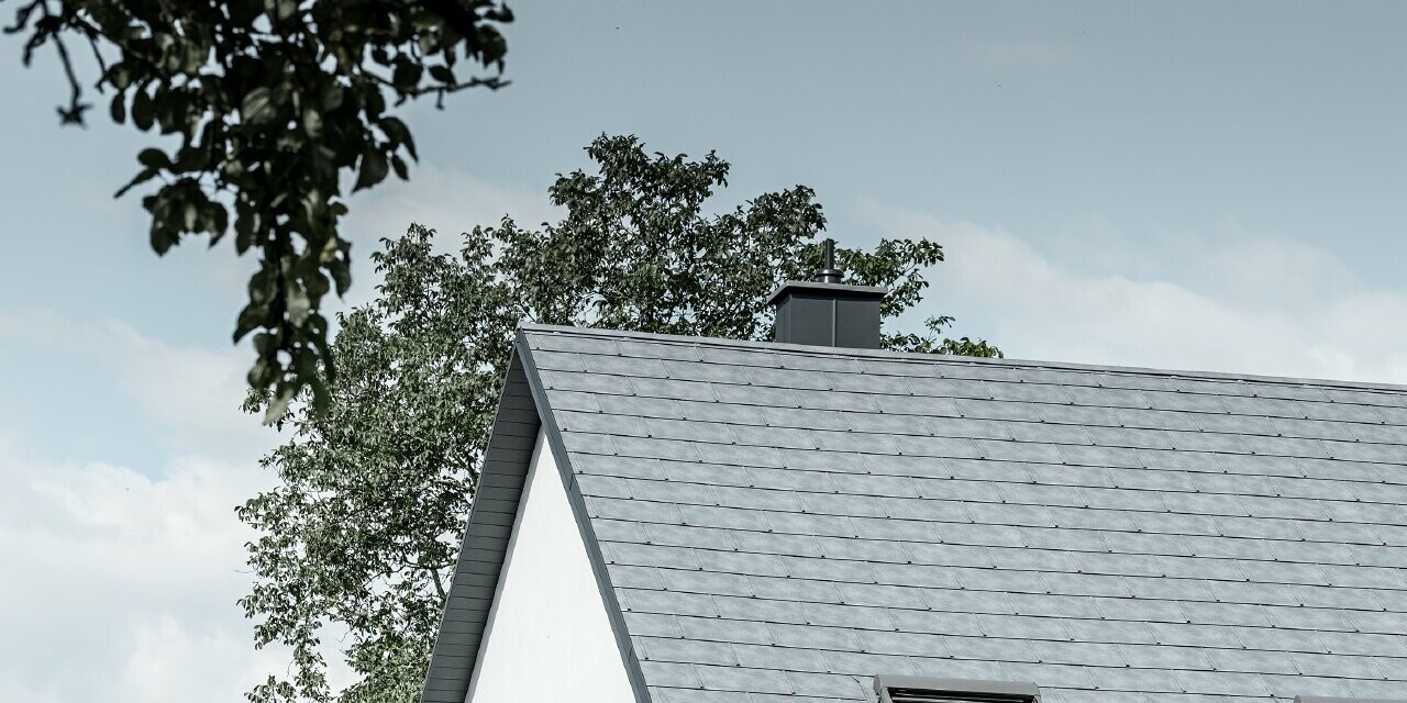 Le toit à deux pans de la maison individuelle classique a été recouvert avec la nouvelle tuile R.16 PREFA couleur gris pierre. Trois fenêtres de toit ont été intégrées dans la surface de toit et des arrêts neige ont été montés. La façade a été simplement laissée blanche.