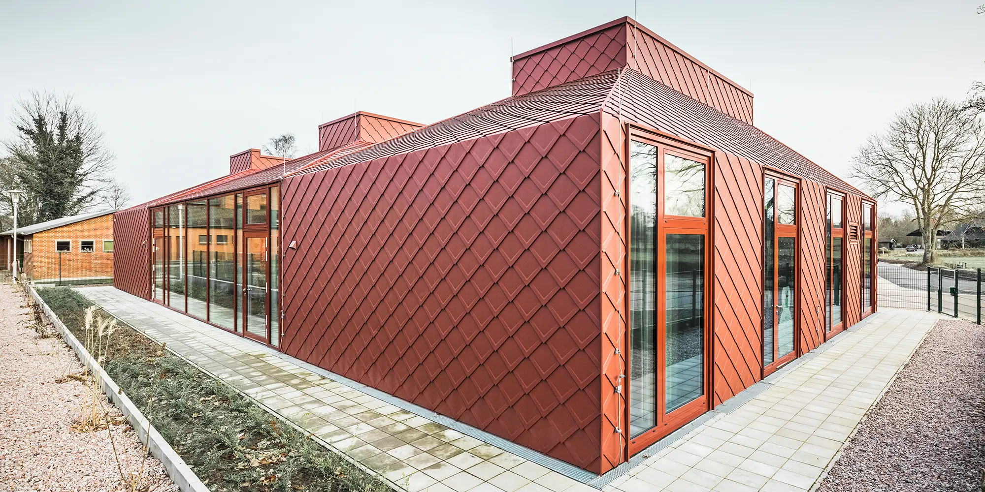 Ein modernes, einstöckiges Gebäude mit einer roten Fassade aus Aluminium, gedeckt mit PREFA Dach- und Wandrauten. Zu sehen sind große, raumhohe Fensterfronten sowie einer klaren, modernen Architektur, fotografiert bei Tageslicht in einer Frontalaufnahme.