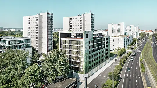 Luftaufnahme des Wohn- und Geschäftshauses „Pick-Nick“ am Straßburger Platz in Dresden mit seiner markanten PREFA-Fassade aus Aluminium-Rhomben in verschiedenen Grün- und Beigetönen. Das Gebäude befindet sich in einem modernen Stadtbild mit Hochhäusern und Grünflächen, direkt an der Hauptstraße. Herausragende Architektur und die hochwertige PREFA-Fassade setzen optische Akzente im Stadtbild.