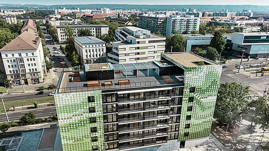 Luftaufnahme des Wohn- und Geschäftshauses „Pick-Nick“ in Dresden mit einer PREFA-Fassade aus Aluminium-Rhomben in verschiedenen Grün- und Beigetönen. Im Hintergrund bietet sich ein beeindruckender Panoramablick über die Stadt. Das Gebäude verfügt über eine großzügige Dachterrasse mit Blumenbeeten und einem Kinderspielplatz. Inmitten urbaner Umgebung vereint es moderne Architektur, Nachhaltigkeit und funktionales Design.