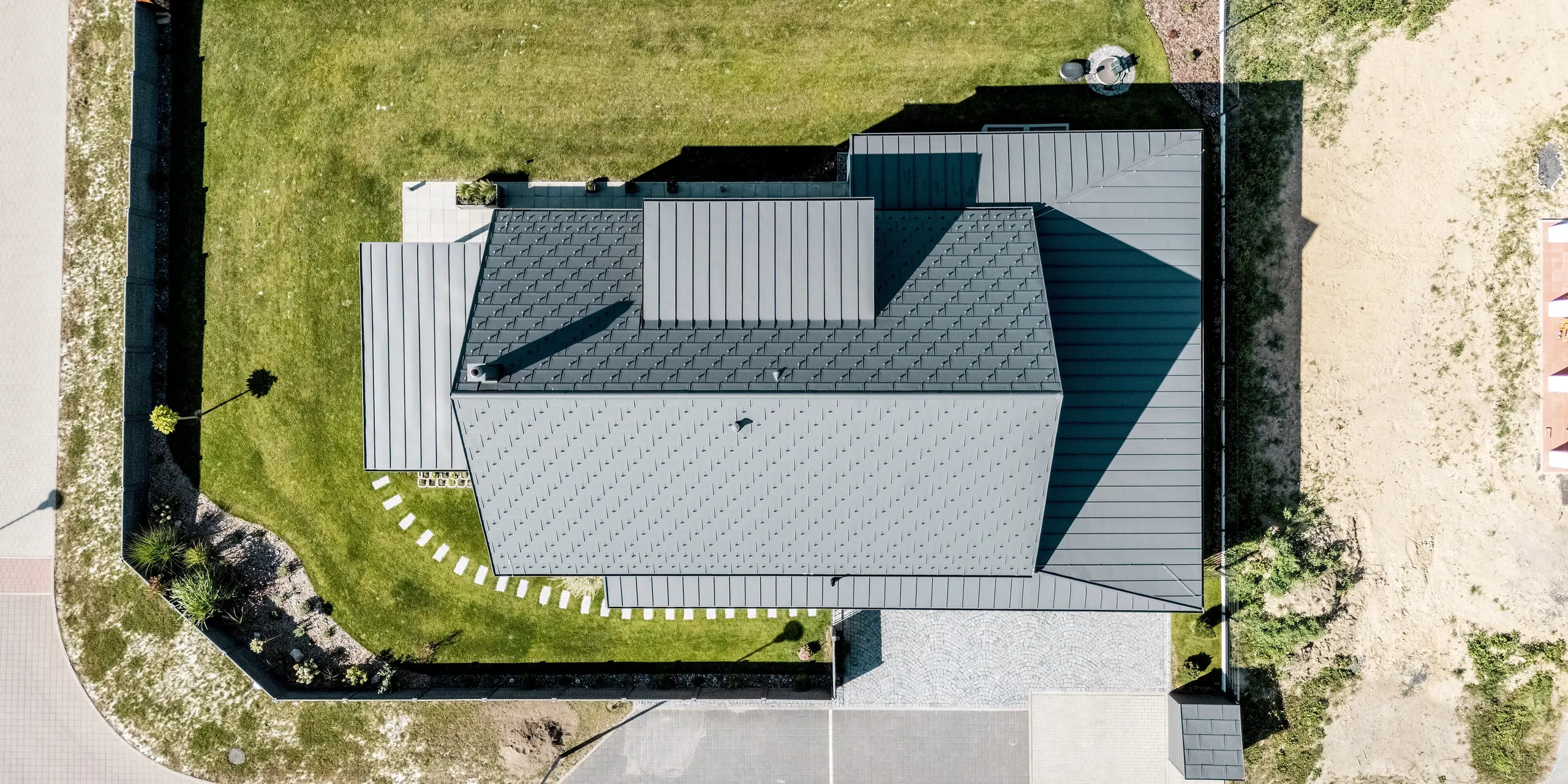 Diese Luftaufnahme zeigt ein modernes Einfamilienhaus in Hlinsko, das mit vielfältigen PREFA Aluminiumlösungen ausgestattet ist. Die klar gegliederten Dachflächen mit PREFA Dachplatten R.16 und PREFALZ in der Farbe P.10 Dunkelgrau zeigen die harmonische Verbindung von Funktion und Design. Die robuste Aluminiumbedachung überzeugt durch Langlebigkeit, Witterungsbeständigkeit und eine elegante, zeitlose Optik. Sie fügt sich perfekt in die gepflegte Gartenanlage ein.