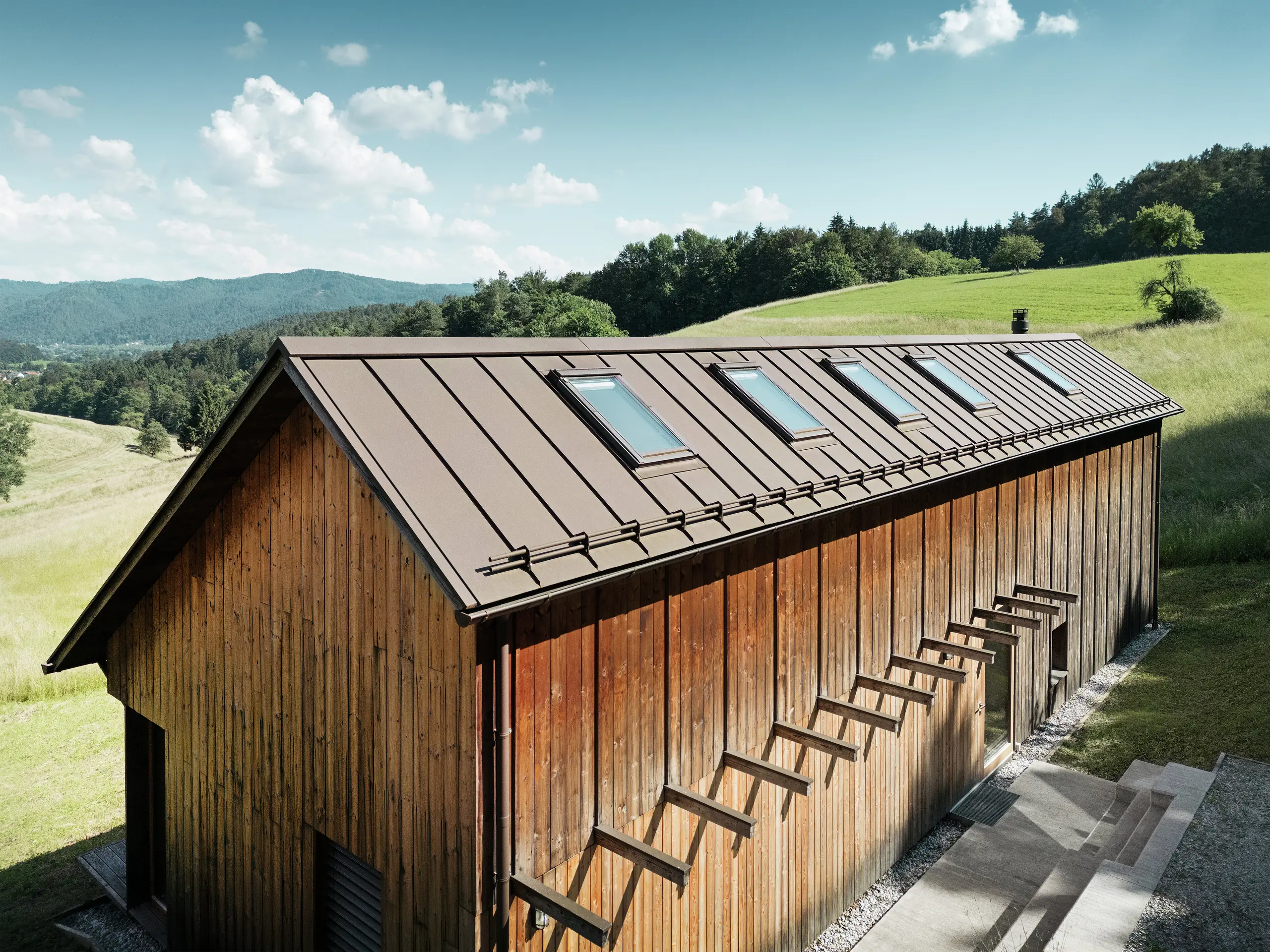 Maison en bois à l’architecture clairement structurée, avec un toit en aluminium PREFALZ de haute qualité en P.10 brun. Ce toit PREFA, nécessitant peu d’entretien, séduit par la précision de ses lignes, sa grande résistance aux UV et à la corrosion, ainsi que par l’intégration parfaite des fenêtres de toit. L’association d’une façade en bois durable et d’une couverture à joint debout longue durée s’intègre harmonieusement dans le paysage vallonné idyllique. Le toit à deux pans asymétrique est équipé de cinq fenêtres de toit posées avec précision, apportant de la lumière à l’intérieur.