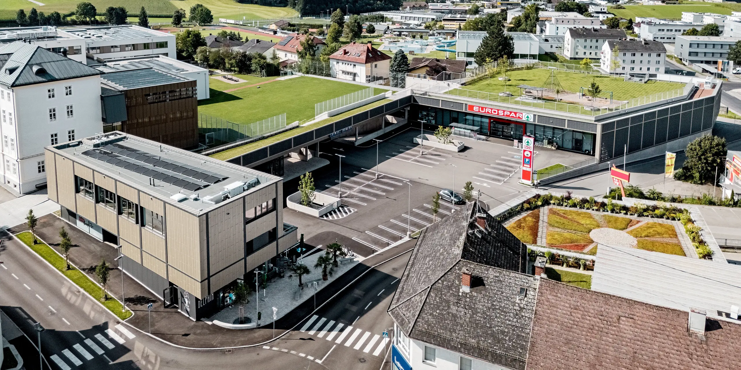 L'impressionnante photographie aérienne du centre commercial de Rohrbach illustre son intégration harmonieuse dans le paysage rural. Au premier plan, le complexe se distingue par sa façade remarquable composée de Siding X PREFA anthracite et bronze. Ces panneaux confèrent à l'ensemble une allure moderne et élégante. Juste en face s'étend un vaste parking, encadré par l'architecture structurée du bâtiment. La pelouse verdoyante et les jardins soignés qui l'entourent créent une atmosphère accueillante. Les maisons alentour et le relief vallonné à l'arrière-plan soulignent la réussite de l'intégration de ce bâtiment moderne dans son environnement.