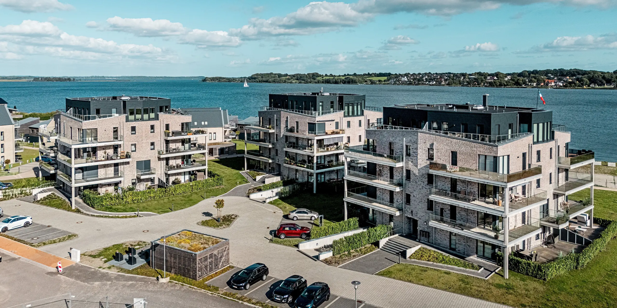 La photographie aérienne présente le complexe résidentiel « Hafen zur Freiheit » (Port vers la liberté) à Schleswig, situé en bord de mer. Il se compose de plusieurs bâtiments modernes aux façades en briques claires, rehaussées de panneaux Siding.X PREFA couleur P.10 anthracite. Ces panneaux structurés en aluminium confèrent aux étages supérieurs une allure contemporaine et dynamique, contrastant nettement avec la maçonnerie traditionnelle. De généreux balcons offrent une vue imprenable sur la mer, tandis que les éléments de façade PREFA, résistants à la corrosion et durables, assurent une protection optimale face aux risques liés à cet environnement côtier et contribuent à la pérennité du complexe.