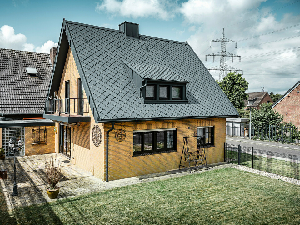 Ein Einfamilienhaus in Tönisvorst mit einem PREFA Dach aus Dachrauten 29×29 in P.10 Anthrazit, das ein herausstechendes Rautenmuster bietet. Schleppgauben, Kamin und der Eingangsbereich sind mit PREFALZ verkleidet, was zusammen mit Ablaufrohr und Hängerinne in P.10 Anthrazit dem Haus einen zeitlosen und eleganten Look verleiht. Das Backsteingebäude mit dem Aluminiumdach harmoniert mit der ruhigen, grünen Umgebung der Siedlung.