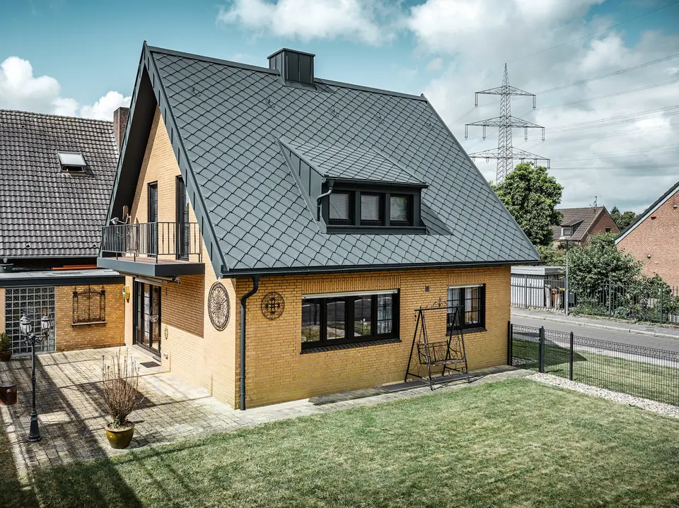 Ein Einfamilienhaus in Tönisvorst mit einem PREFA Dach aus Dachrauten 29×29 in P.10 Anthrazit, das ein herausstechendes Rautenmuster bietet. Schleppgauben, Kamin und der Eingangsbereich sind mit PREFALZ verkleidet, was zusammen mit Ablaufrohr und Hängerinne in P.10 Anthrazit dem Haus einen zeitlosen und eleganten Look verleiht. Das Backsteingebäude mit dem Aluminiumdach harmoniert mit der ruhigen, grünen Umgebung der Siedlung.