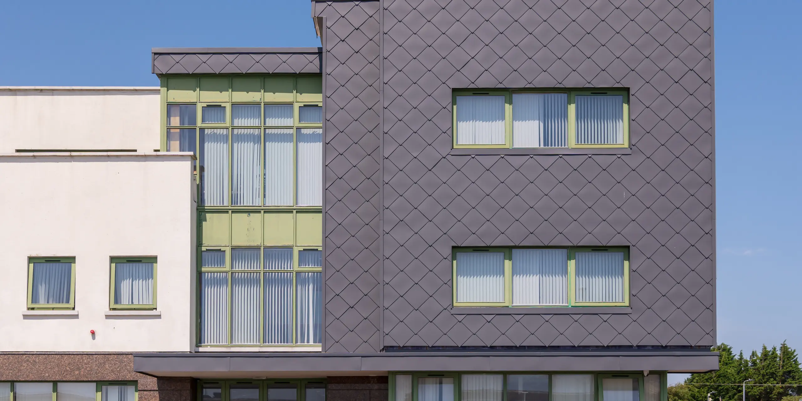 Gros plan dynamique de la façade du centre de santé mentale pour enfants et adolescents de Wexford, revêtue de losanges 44 x 44 PREFA  en P.10 gris sombre : la structure précise des losanges crée de magnifiques jeux d’ombre et de lumière, tandis que les fenêtres à cadre vert apportent du contraste. La robuste façade en aluminium impressionne par sa durabilité, son faible entretien et sa grande résistance aux intempéries.