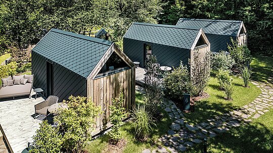 Vue aérienne de plusieurs mini-maisons minimalistes du Secret Koi Garden, au bord du lac Natterer, nichées au cœur d'une végétation luxuriante et de sentiers de jardin naturels. Toits et façades sont entièrement revêtus de losanges en aluminium PREFA P.10 anthracite, dont la texture uniforme en écailles de poisson définit l'esthétique générale. Léger, durable et résistant aux intempéries, l'aluminium PREFA permet une grande précision dans les détails et souligne les lignes épurées de cette architecture de glamping, en parfaite harmonie avec la nature.