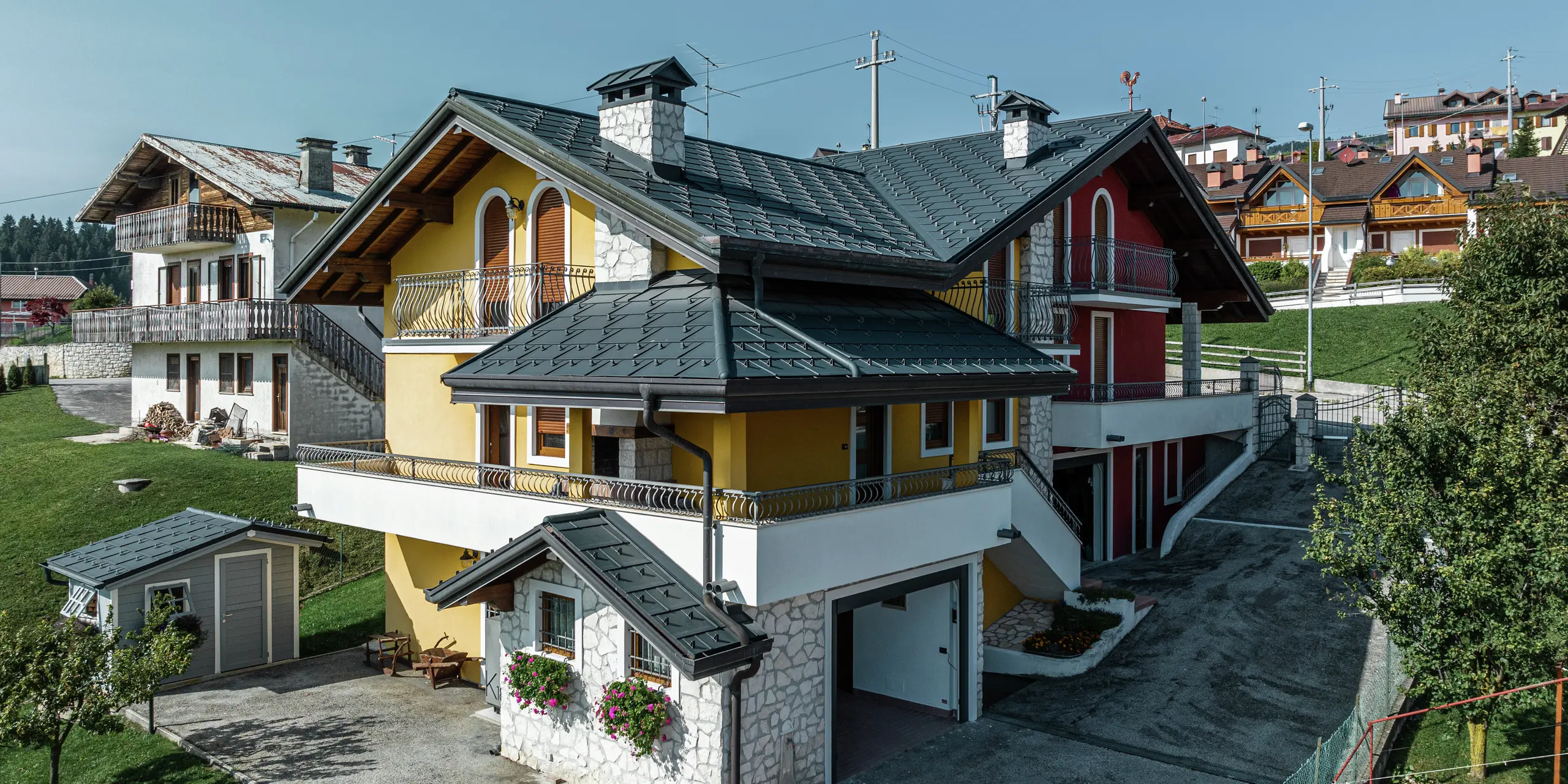 Maison individuelle à Gallio de style architectural alpin avec toiture moderne en R.16 PREFA en P.10 gris sombre. Les panneaux de toiture PREFA garantissent une solution de toiture durable, résistante aux intempéries et visuellement attrayante. La combinaison harmonieuse de la façade jaune-rouge, des éléments en pierre naturelle et des accents en bois confère à la maison un caractère accueillant. Le système de gouttière PREFA en P.10 gris sombre assure un drainage fiable de l'eau. Un excellent exemple d’architecture esthétique et fonctionnelle dans un environnement alpin.