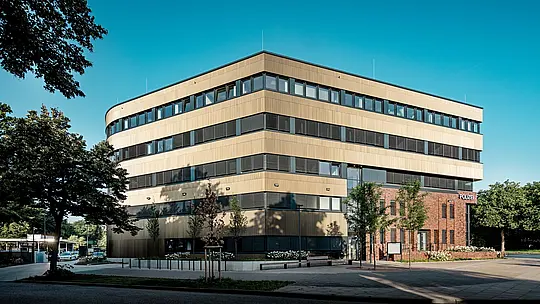 Vue extérieure panoramique du commissariat 43 à Hambourg : le nouveau bâtiment, à la structure affirmée, avec ses angles arrondis et ses bandes de fenêtres horizontales, impressionne par sa façade en aluminium PREFA d’environ 1 100 m², composée de profils extrudés « triangle » couleur bronze moyen. La surface finement texturée révèle des reflets vibrants selon la lumière et confère au bâtiment profondeur et élégance. Le contraste saisissant avec la maçonnerie existante crée un point focal remarquable, tandis que l’aluminium PREFA offre durabilité, résistance à la corrosion et facilité d’entretien.