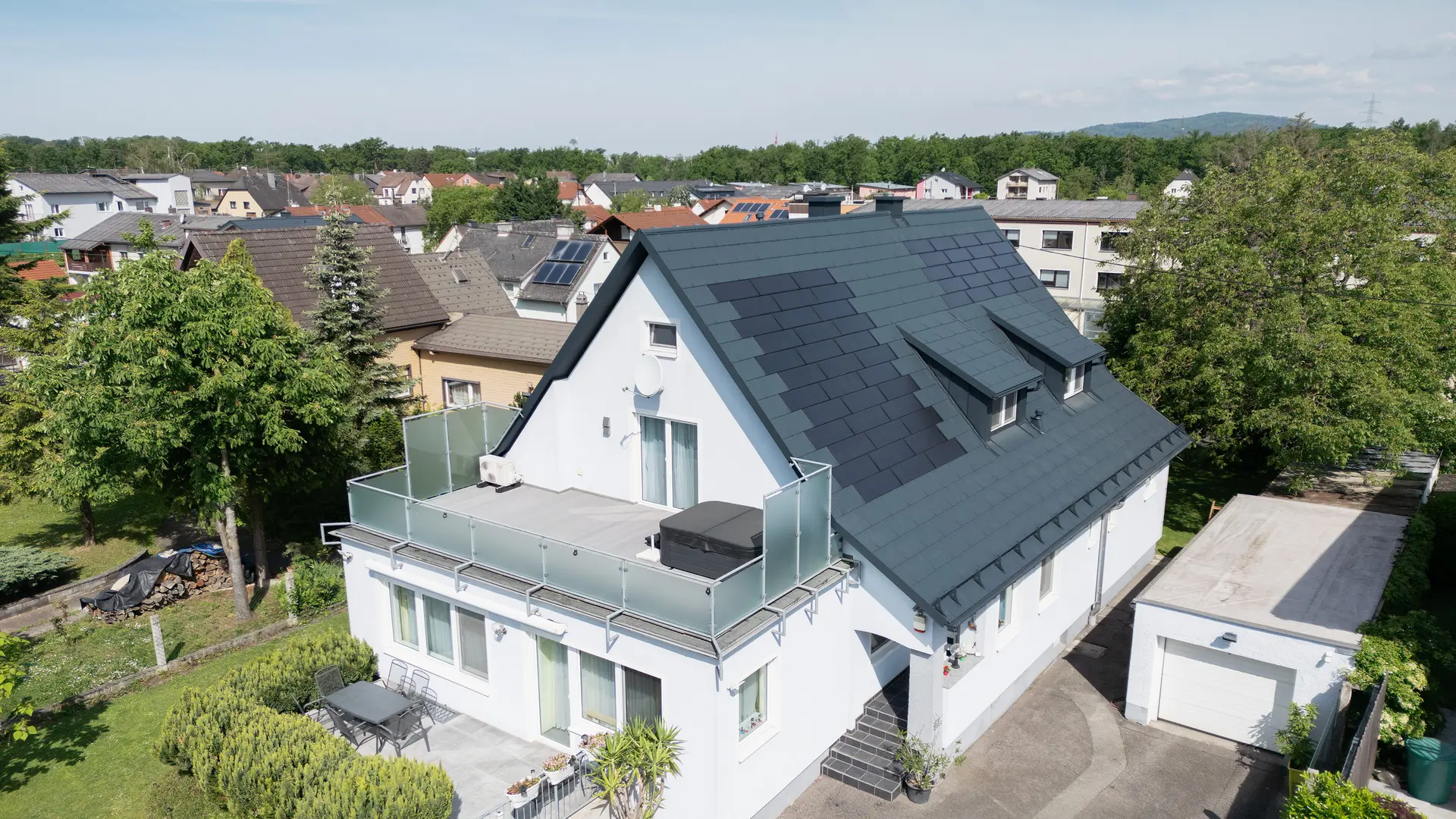 Vue aérienne d'une maison individuelle après la rénovation de la toiture. Le toit est désormais recouvert de grandes tuiles solaires PREFA et de tuiles PREFA R.16 en P.10 anthracite. Les tuiles solaires sont réparties de manière homogène et offrent une solution moderne et durable. Le bâtiment présente désormais un aspect moderne et respectueux de l'environnement. Le balcon au premier plan est doté d'une balustrade en verre et se trouve à l'étage supérieur du bâtiment.