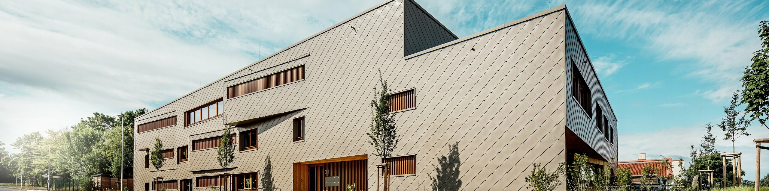 Vue en contre-plongée, la crèche d'Alzenau se distingue par ses lignes dynamiques et sa façade en aluminium PREFA bronze de haute qualité. La disposition diagonale des losanges de façade 44x44 confère au bâtiment une structure claire et une surface élégante et homogène. Des ouvertures de fenêtres décalées et des persiennes verticales en bois créent des accents rythmiques, tandis que le design minimaliste traduit une sobriété moderne. L'alliance de l'aluminium durable, du bois naturel et d'un concept d'aménagement paysager bien pensé le long de la rue souligne les exigences élevées en matière de durabilité et de qualité de construction. Cette image illustre avec brio comment une architecture fonctionnelle utilisant les produits PREFA peut être esthétiquement séduisante et définir de nouvelles normes en matière d'urbanisme.