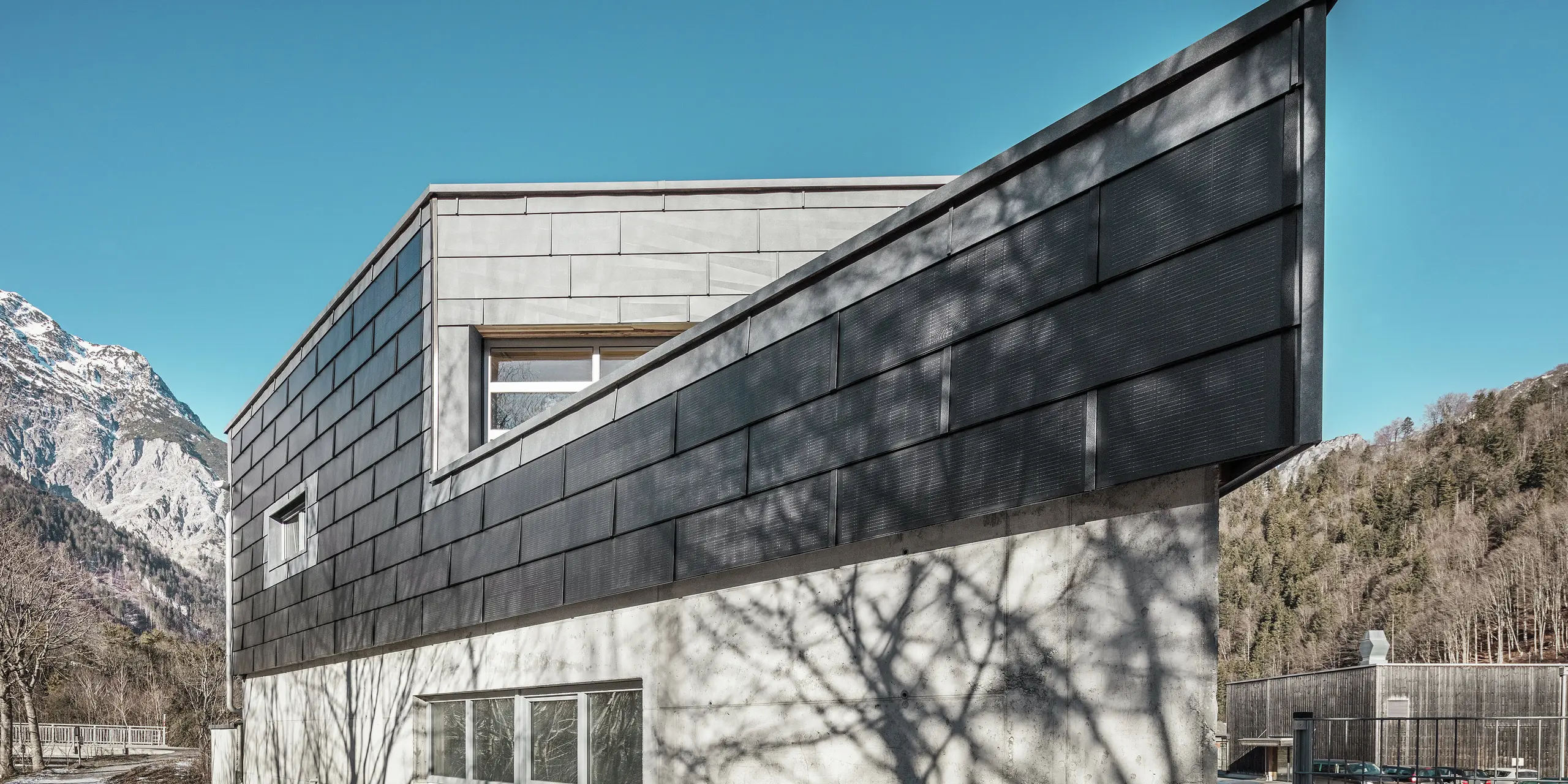 Le bâtiment de l'entreprise métallurgique RÜMA, dans le Vorarlberg, impressionne par sa façade moderne en tuiles solaires PREFA et béton apparent. Entouré de montagnes et d'arbres, dont les ombres se projettent sur le mur, il se dresse sous un ciel bleu et dégagé. L'angle pointu du bâtiment et les panneaux de façade PREFA FX.12 soulignent encore son allure imposante.