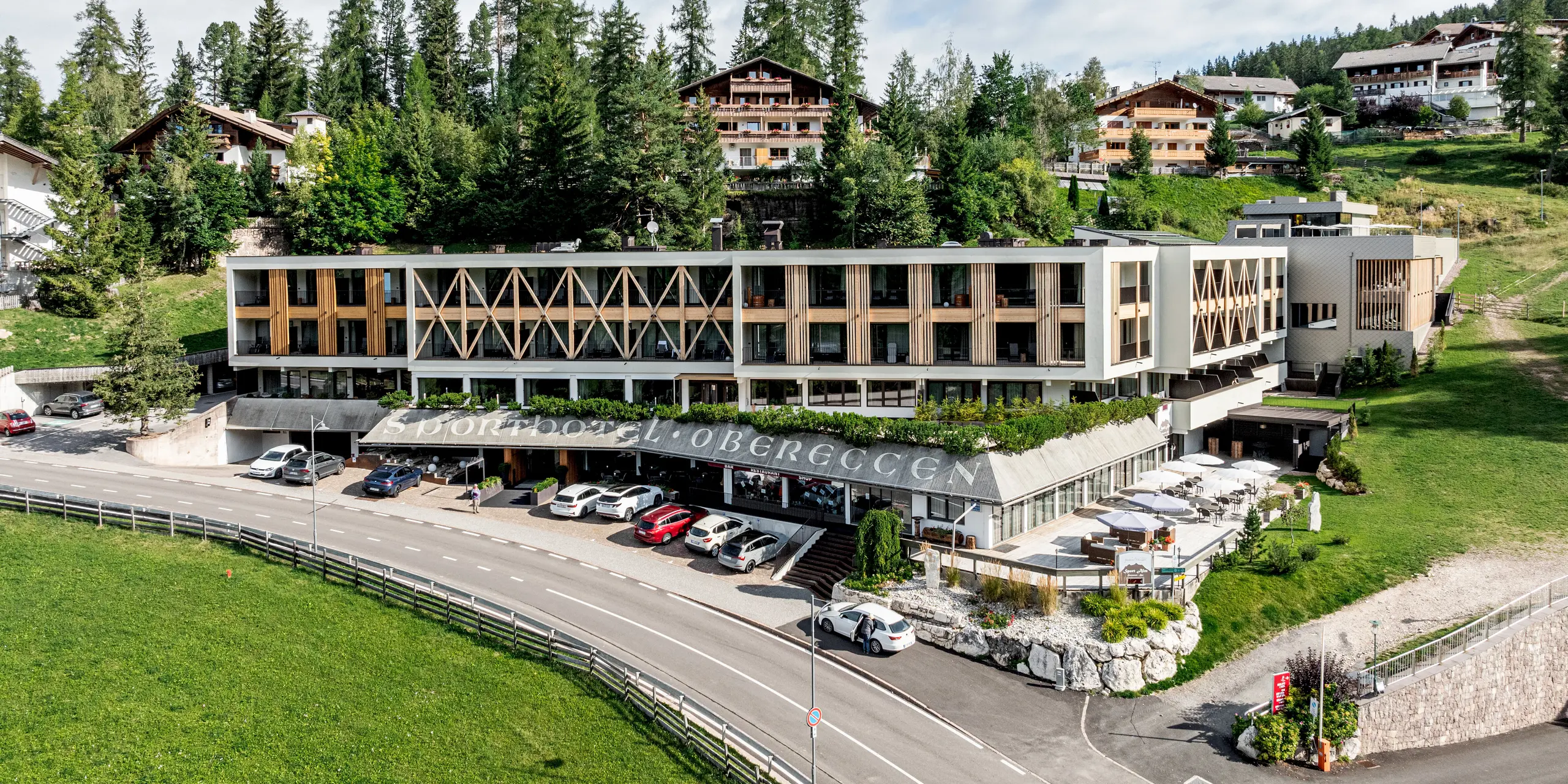 Vue d'ensemble du Sporthotel Obereggen, au Tyrol du Sud, harmonieusement intégré au paysage alpin, entre prairies, arbres et bâtiments à flanc de colline. L'architecture moderne de l'hôtel se caractérise par une façade en aluminium PREFA composée de losanges 29x29 couleur bronze, qui confère au bâtiment structure, profondeur et élégance. La chaude teinte bronze crée un contraste saisissant avec le bois et l'environnement naturel. Durables, résistants aux intempéries et nécessitant peu d'entretien, les produits en aluminium PREFA sont parfaitement adaptés à une architecture haut de gamme en régions alpines.