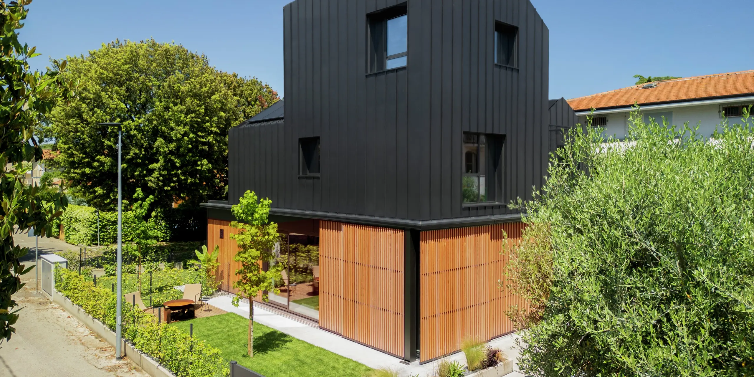 Vue de rue d'une maison jumelée moderne à Cattolica : une élégante combinaison de lattes de bois au rez-de-chaussée et d'une façade en aluminium PREFALZ gris noir. Les lignes épurées et la surface de haute qualité PREFA offrent durabilité, protection contre la corrosion et une architecture intemporelle sous un ciel ensoleillé.