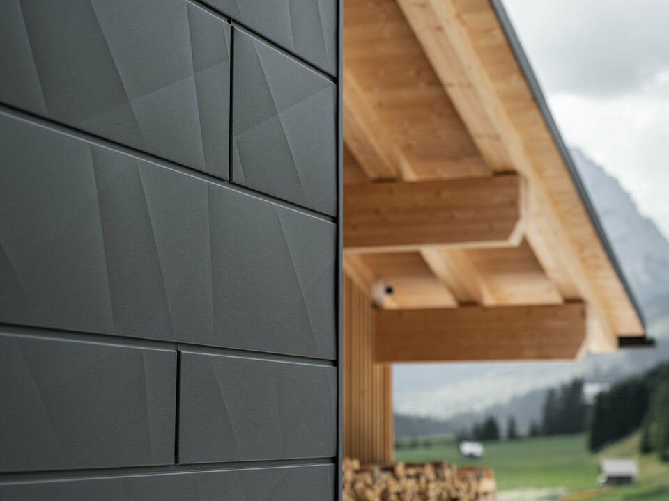 Gros plan sur les panneaux de façade Siding.X en P.10 noir de PREFA sur la Casa Hoffe à Sappada, en Italie. Le système de façade en aluminium est habilement intégré dans un design moderne et abstrait qui contraste agréablement avec les éléments en bois apparents de la charpente de toit. Cette solution de façade innovante s'intègre parfaitement dans l'environnement alpin et allie fonctionnalité, durabilité et esthétique architecturale.