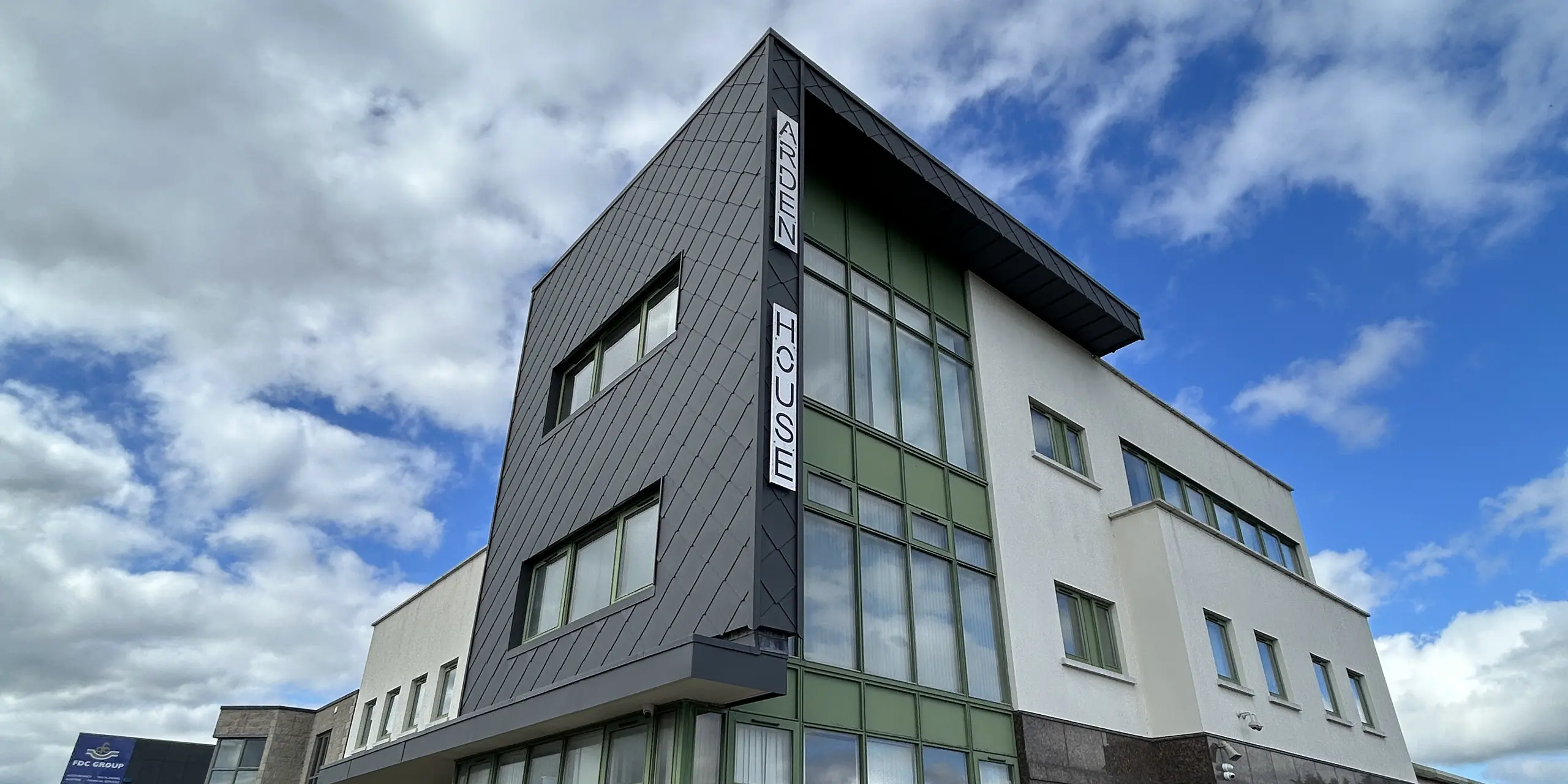 Markanter Gebäudekubus des Arden House, Zentrum für psychische Gesundheit von Kindern und Jugendlichen, in Wexford mit PREFA Wandraute 44x44 in P.10 Dunkelgrau. Die dynamische Rautenstruktur betont die klare Architektur, während die grünen Fenster Akzente setzen. Die langlebige und korrosionsbeständige Aluminiumfassade überzeugt durch Wartungsarmut und hohe Widerstandsfähigkeit.