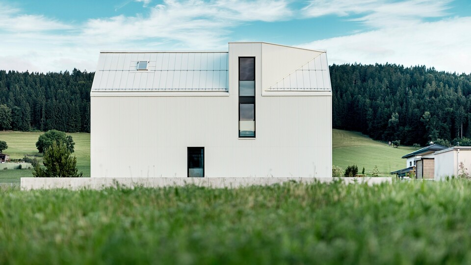 L'image présente une maison individuelle à Freistadt, vue de profil, mettant en valeur son architecture minimaliste et remarquable. La façade en aluminium blanc pur PREFALZ et le toit à pignon abrupt dessinent une silhouette épurée et moderne qui s'intègre harmonieusement à la verdure environnante. Les grandes fenêtres et l'élégante lucarne soulignent la fonctionnalité du design, tandis que la qualité de l'aluminium garantit à la fois une esthétique durable et une excellente résistance aux intempéries. Par sa forme simple et l'utilisation de matériaux pérennes, le bâtiment dégage une élégance intemporelle et représente une parfaite harmonie entre architecture et nature.