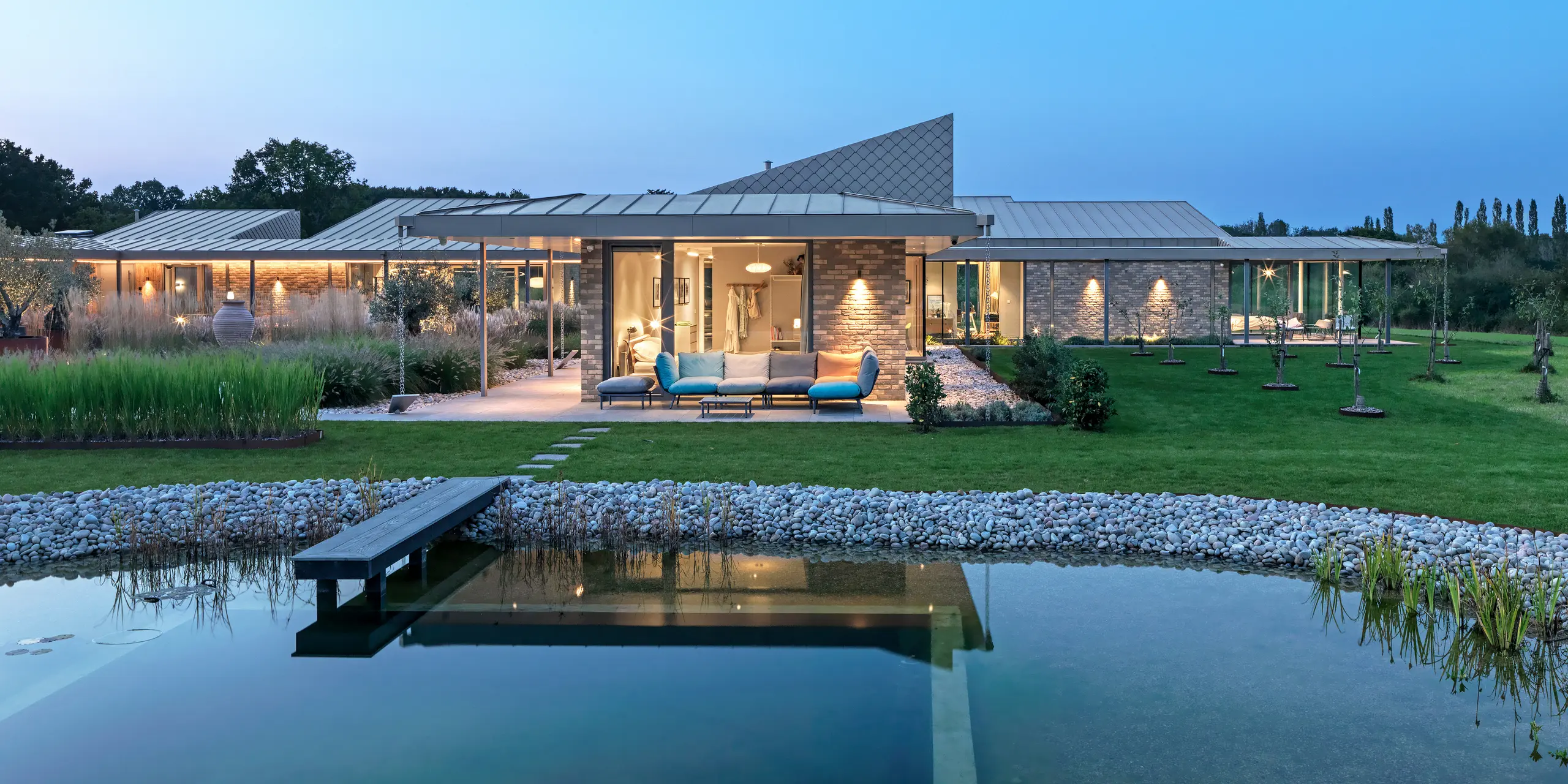 Vue nocturne d'une magnifique maison du West Sussex, avec toiture en aluminium PREFALZ et losanges de façade PREFA 20 × 20 P.10 bronze. Au premier plan, une piscine naturelle paysagée avec une passerelle en bois, derrière laquelle se trouve une spacieuse terrasse avec mobilier de jardin et une véranda éclairée. Les lignes épurées de l'architecture et la qualité des produits PREFA créent une élégante alliance de design, de fonctionnalité et de durabilité.