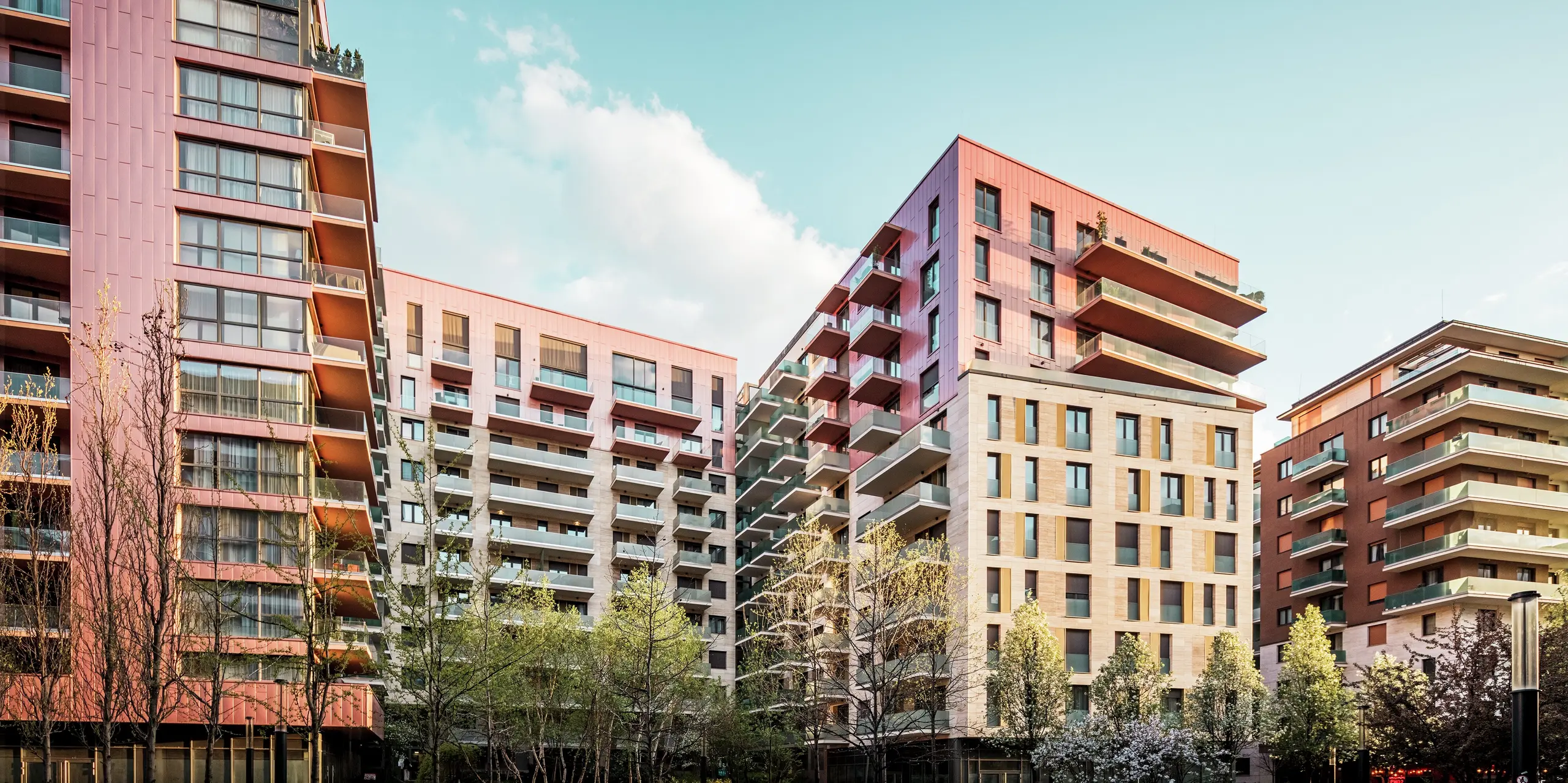 Vue d'ensemble du projet résidentiel Grand Corvin, dans le quartier revitalisé de Corvin à Budapest. Le revêtement de façade Siding PREFA, d'une couleur saumon métallisée, crée des accents visuels forts et met en valeur les étages supérieurs. Associé à la pierre naturelle et au verre, il crée un design architectural moderne et de haute qualité. Les panneaux en aluminium durables sont faciles d'entretien, résistants aux intempéries et s'intègrent harmonieusement à l'environnement urbain. La photo a été prise à l'heure dorée, ce qui permet à la lumière agréable et chaleureuse d'encadrer la scène.