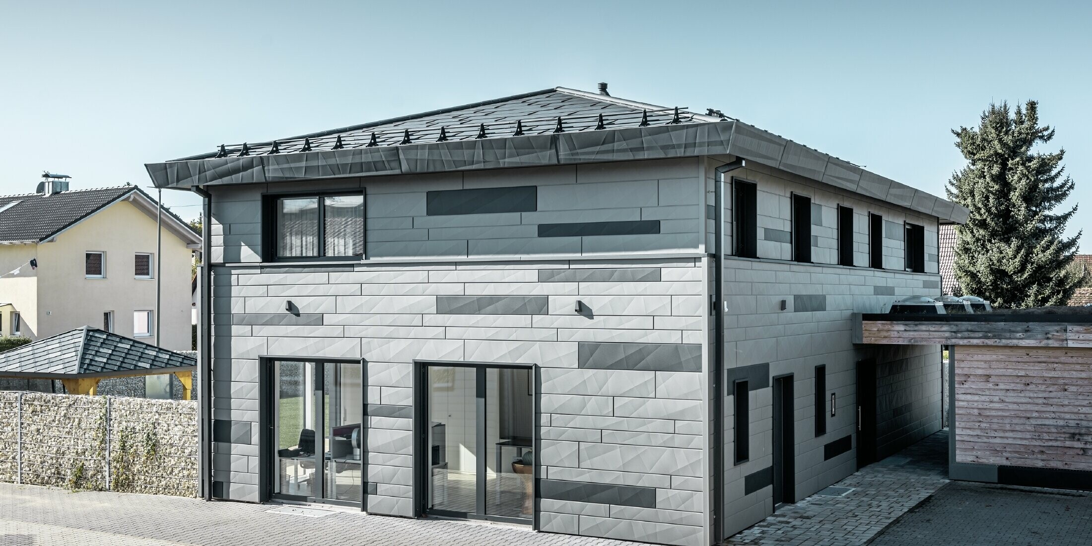 Vue sur la maison individuelle depuis la rue. La façade bicolore et les arêtes des panneaux FX.12 donnent une apparence moderne à la maison.