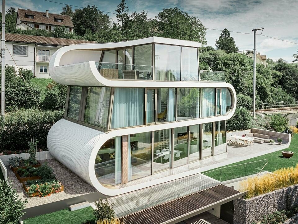 Photo de la Flexhouse moderne au bord du lac de Zurich. Avec sa forme hors du commun, la Flexhouse attire toujours le regard. Les séparations entre étages s’étirent comme une bande blanche ondulée à travers le bâtiment. Les éléments de façade arrondis ont été recouverts de bardeaux PREFA en aluminium blanc pur.