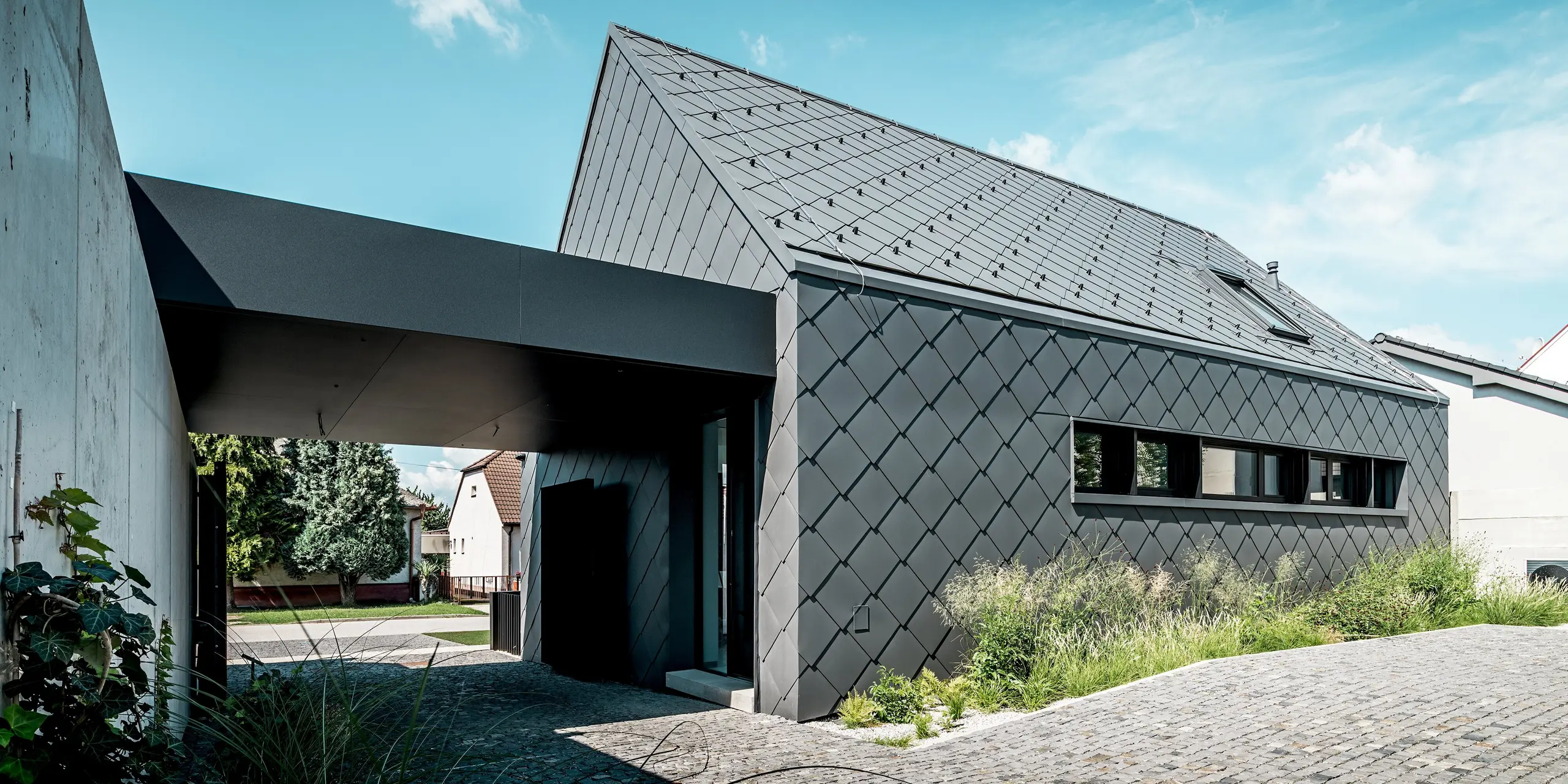 L'image présente l'architecture moderne d'une maison individuelle à Trnava-Modranka, entièrement revêtue de losanges de toiture et de façade 44x44 PREFA en colorisP.10 gris sombre. La structure continue en losanges souligne la pureté des lignes du bâtiment et lui confère une allure élégante et monolithique. Ces produits en aluminium résistant assurent une protection durable contre les intempéries et une enveloppe de bâtiment de haute qualité, nécessitant peu d'entretien. La façade avant, baignée de lumière, et la végétation environnante créent un contraste naturel avec l'esthétique remarquable des panneaux PREFA.