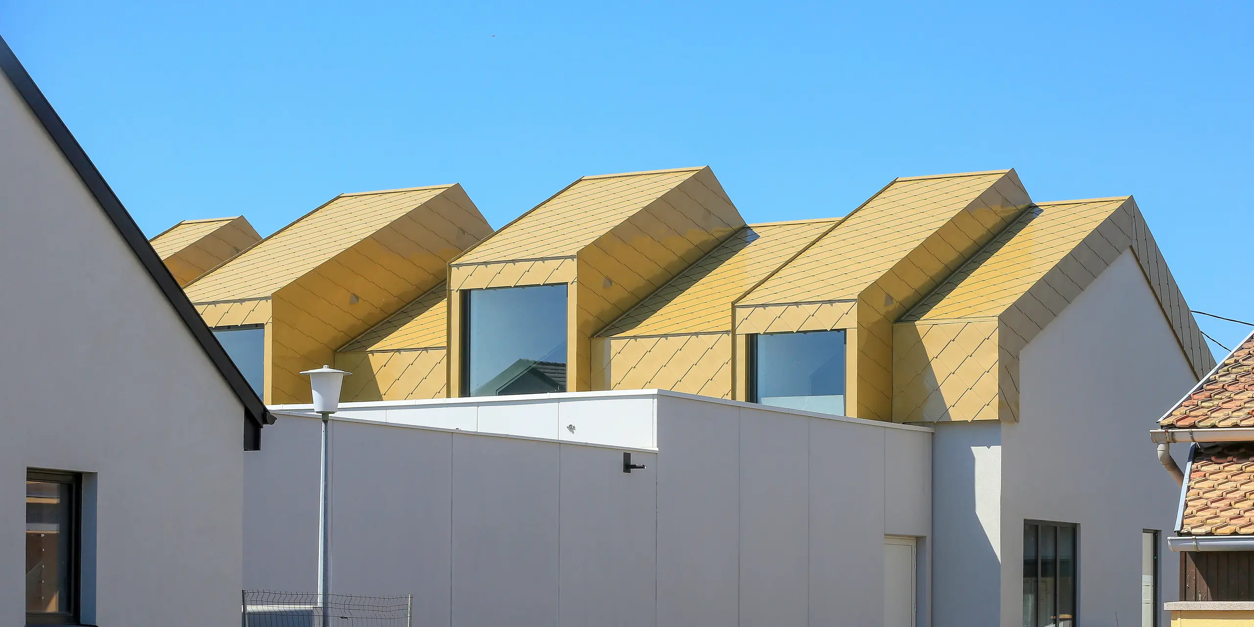 Vue de loin du Centre Communautaire et Culturel de Fessenheim (France). La perspective du bâtiment avec le toit brillant PREFA et les losanges de façade 44 × 44 en Sparkling Gold est depuis la rue.