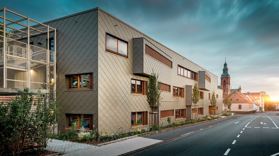 Sous la douce lumière du crépuscule, la crèche d'Alzenau révèle toute sa splendeur. Sa façade, revêtue de losanges de façade PREFA couleur bronze (44 x 44 cm), reflète la lumière du soir et met en valeur la texture raffinée de ce bâtiment hybride et moderne. L'architecture aux lignes épurées, avec ses bandes de fenêtres décalées et ses persiennes verticales en bois, contraste avec l'église historique qui se dresse en arrière-plan. À gauche, une délicate structure métallique apporte une touche de transparence à la cage d'escalier extérieure. La bande verte soigneusement entretenue qui longe le bâtiment témoigne de son engagement en faveur du développement durable. Cette image illustre avec brio la combinaison d'une architecture moderne, de la technologie durable de l'aluminium PREFA et d'une intégration harmonieuse au paysage urbain historique d'Alzenau.