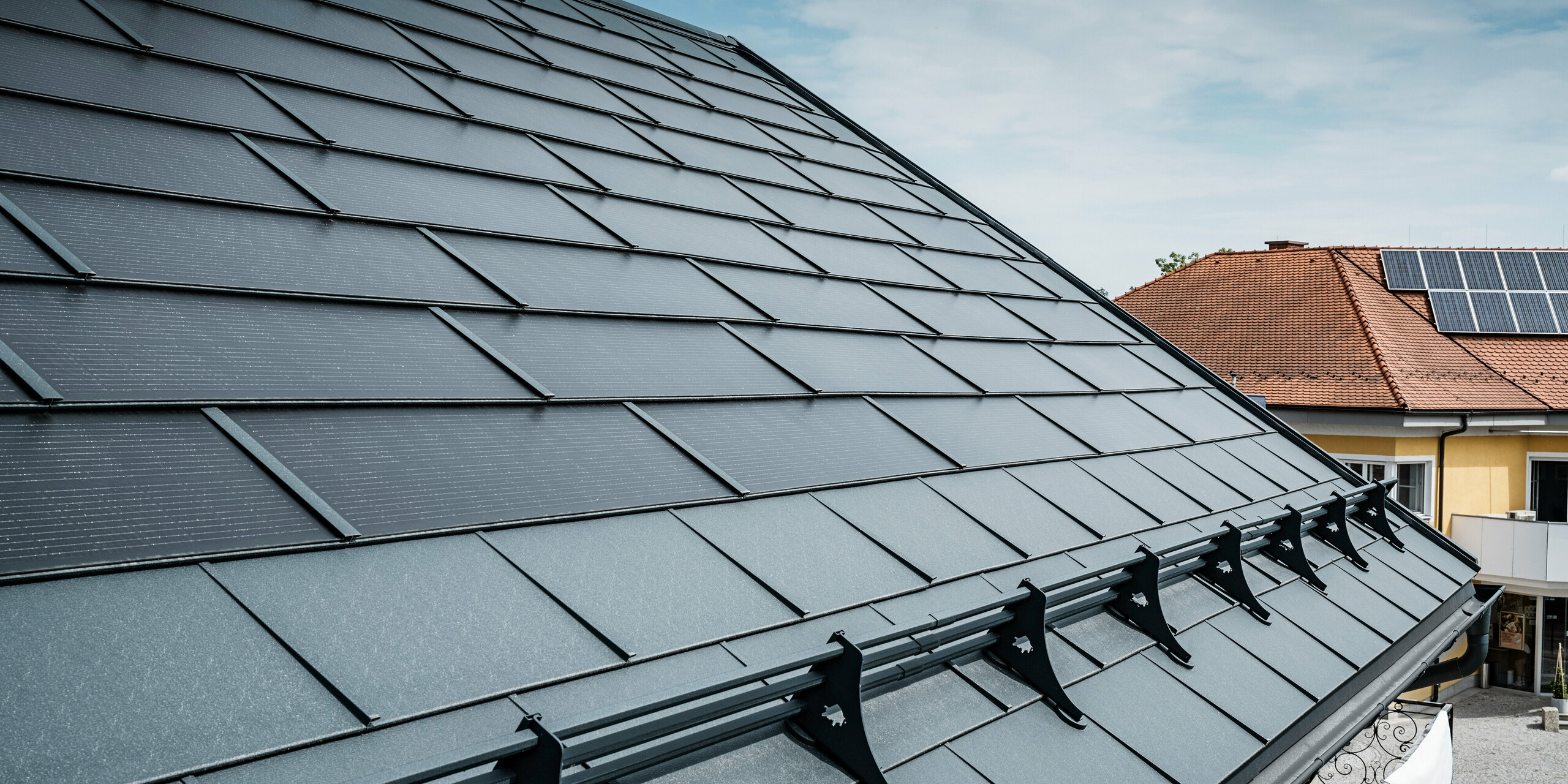 Ansicht eines modernen Daches mit anthrazitfarbenen PREFA Solardachplatten. In der Nähe der Traufe wurde das Dach mit klassischen PREFA Dachplatten R.16 und einem PREFA Schneeschutzsystem ausgestattet. Im Hintergrund ist das Dach eines benachbarten Hauses mit traditionellen Dachziegeln und Aufdach-Solarmodulen zu sehen, was den Kontrast zwischen neuer und herkömmlicher Dachgestaltung hervorhebt. Die innovative Solardachplatte von PREFA vereint Ästhetik und Funktionalität und bietet eine nachhaltige Energielösung für alle Gebäudearten.