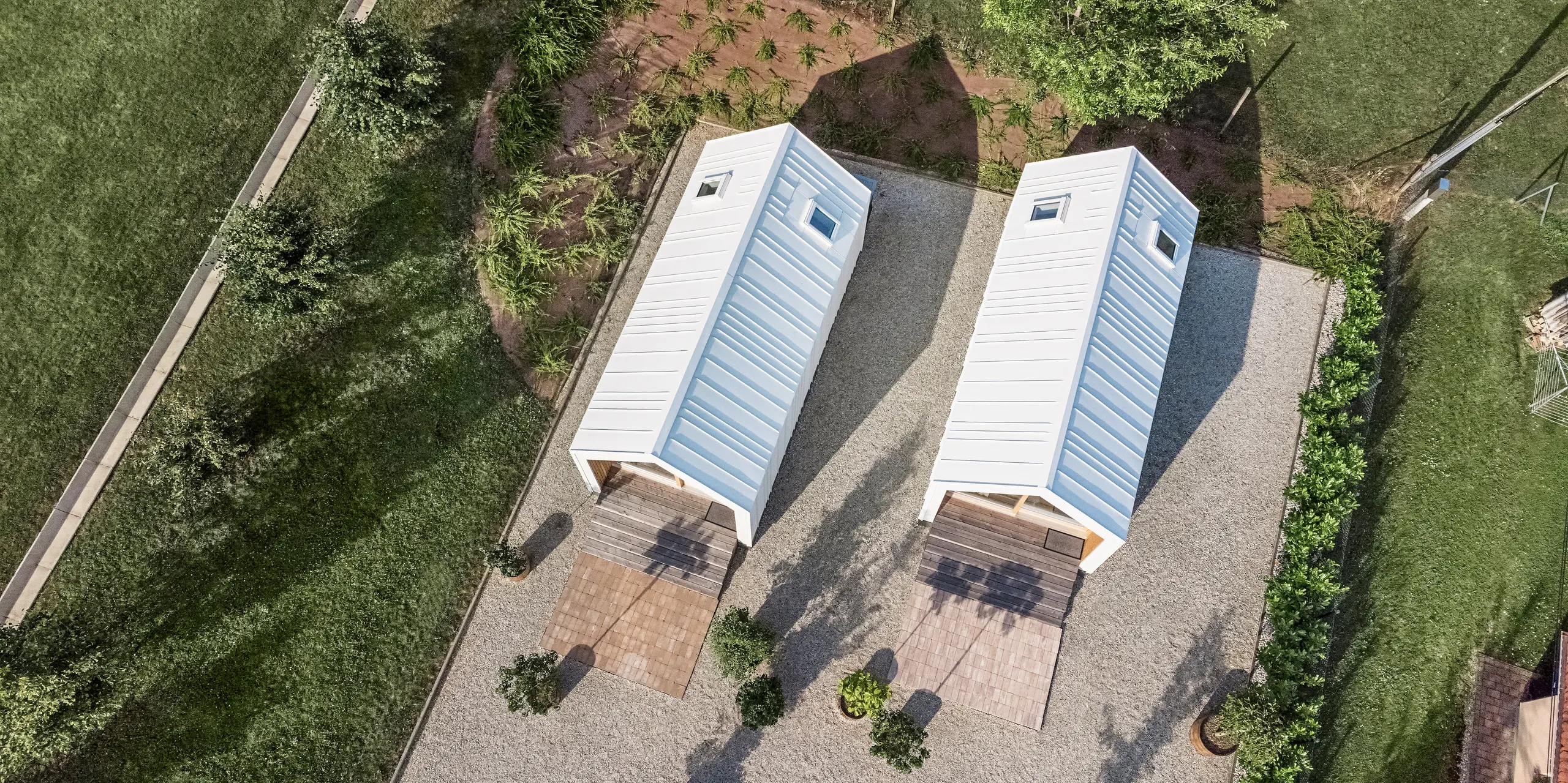 Vue aérienne des deux petites maisons du complexe de vacances « Pri Momi » à Kančevci, en Slovénie, conçues avec un revêtement de toit et de façade PREFALZ en blanc pur P.10. La photo montre la disposition symétrique des deux bâtiments avec leur coque blanche en aluminium robuste de haute qualité et les espaces verts environnants. Les lignes claires et l'architecture moderne des maisons sont soulignées par l'exécution précise du revêtement en aluminium. Les sentiers environnants et l'environnement naturel s'intègrent harmonieusement dans l'ensemble et invitent les visiteurs à s'attarder.
