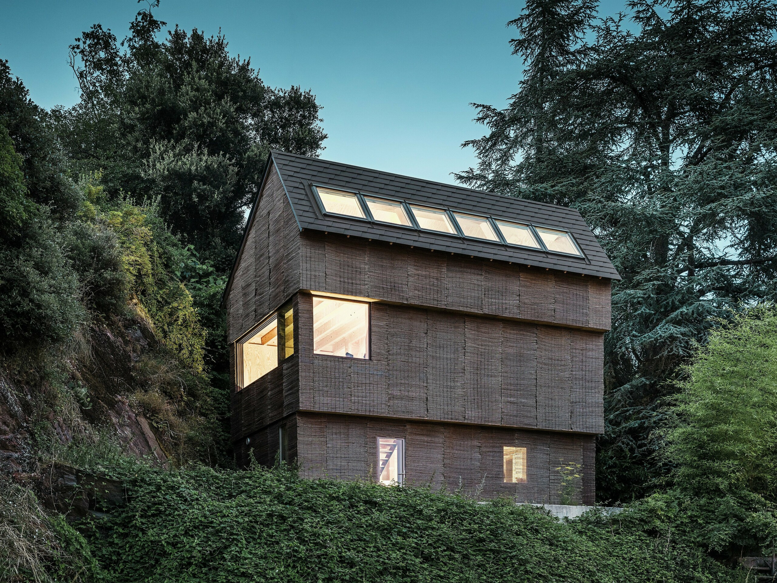 Vue nocturne d'une maison moderne dans l'ouest de la France avec des bardeaux de toiture PREFA en P.10 brun noisette, des puits de lumière spacieux et une façade en bois de saule durable.