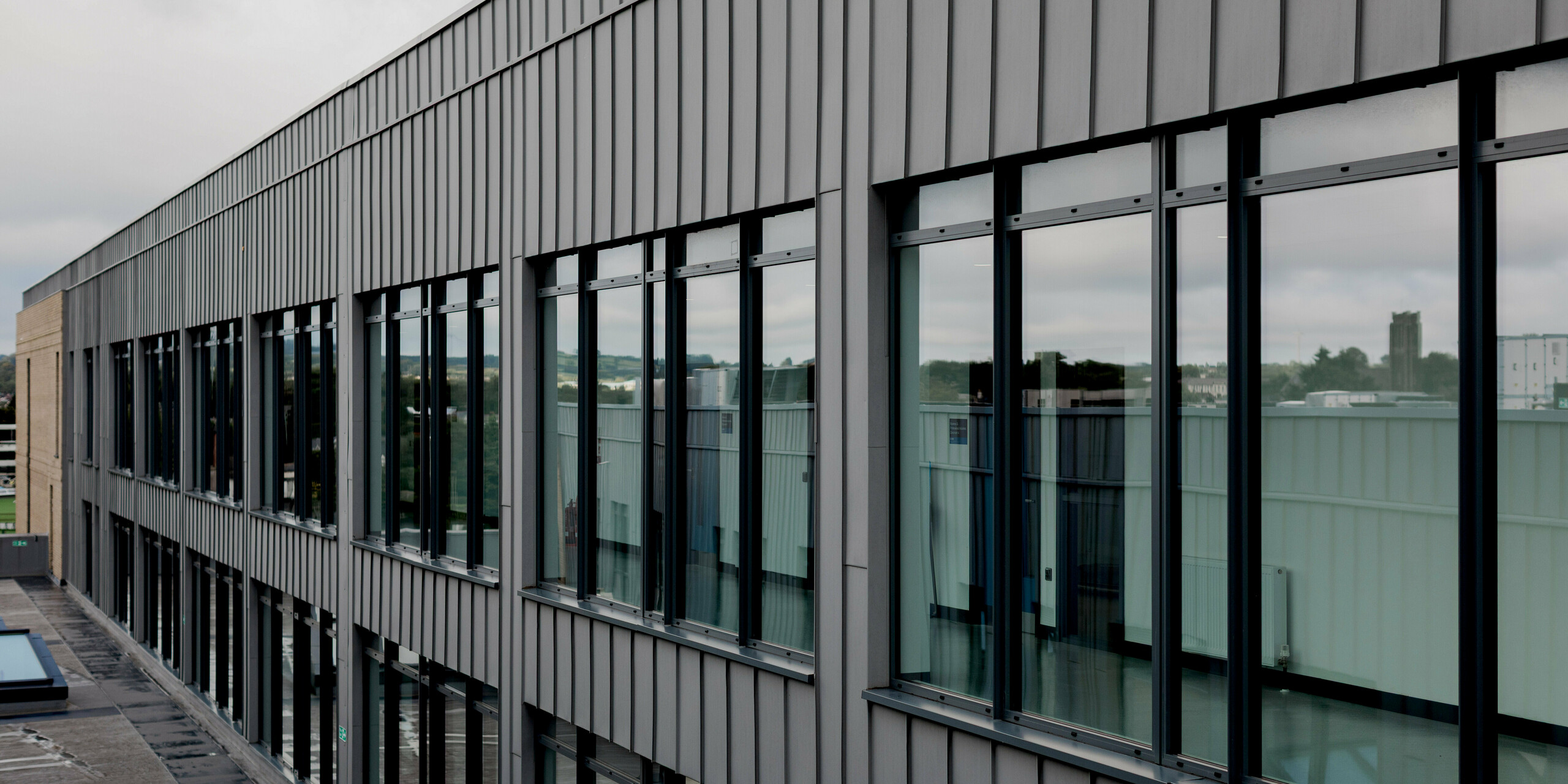 Eine langgestreckte Fassadenansicht des Northern Regional College in Ballymena zeigt eine durchgehende PREFALZ Aluminiumfassade in Patinagrau mit streng vertikaler Stehfalzstruktur. Große, gleichmäßig gereihte Fensterflächen spiegeln den Himmel und die Umgebung und verleihen dem Gebäude Transparenz und Tiefe. Die metallische Hülle bildet einen ruhigen, klaren Rahmen für die Glasflächen und unterstreicht die lineare Architektur. PREFA Aluminium überzeugt hier durch Präzision, Langlebigkeit, Witterungsbeständigkeit und eine hochwertige, moderne Optik.