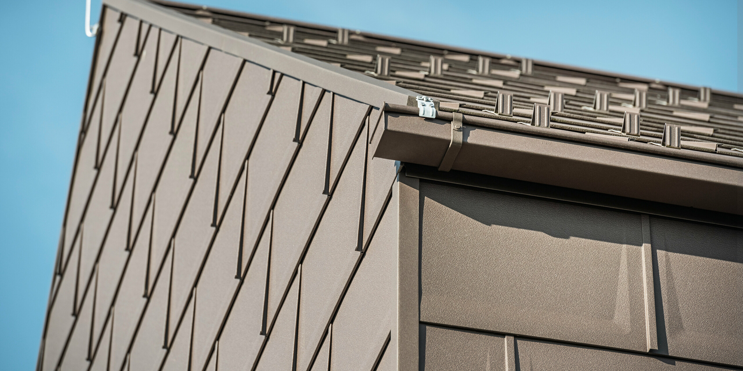 Detailaufnahme der Dachentwässerung eines Einfamilienhauses in Neukirchen, Österreich, mit einer PREFA Kastenrinne. Die Dachplatte R.16 in P.10 Braun bildet nicht nur das Dach, sondern ziert auch die Fassade, was ein durchgängiges und elegantes Erscheinungsbild schafft. Diese Nahaufnahme zeigt die Präzision und Qualität der PREFA-Aluminiumprodukte, die Design mit Funktionalität für ein modernes Wohnkonzept verbinden.