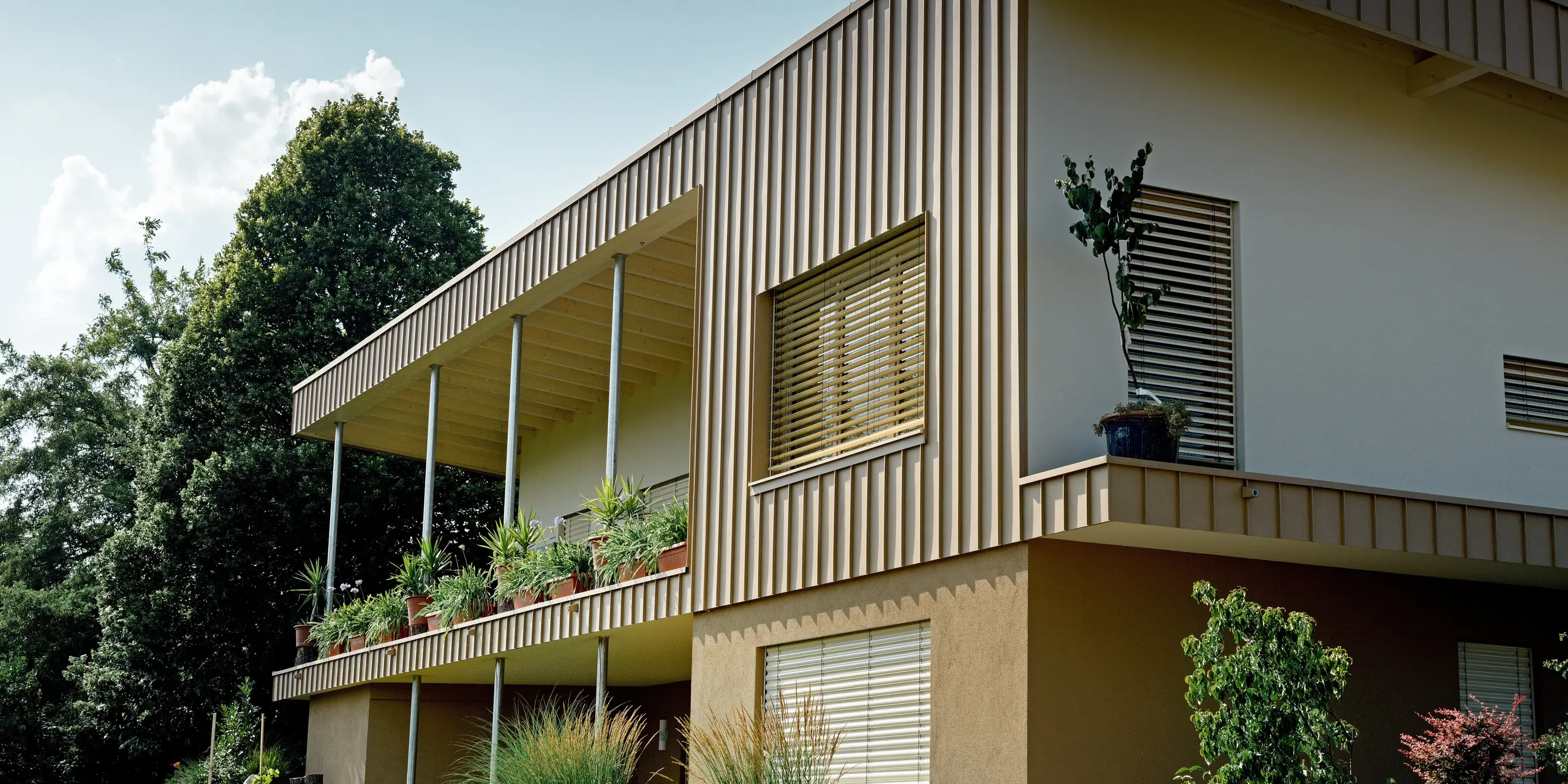 Perspektivische Ansicht des Einfamilienhauses in Schiefling am See mit markanter PREFALZ Fassade aus Aluminium in P.10 Sandbraun. Die prägnanten Falzprofile rahmen die Architektur eindrucksvoll ein und lenken den Blick auf die großzügige Terrasse mit üppiger Begrünung. Die langlebige PREFA Aluminiumfassade überzeugt durch zeitlose Eleganz, Wartungsarmut und hohe Witterungsbeständigkeit