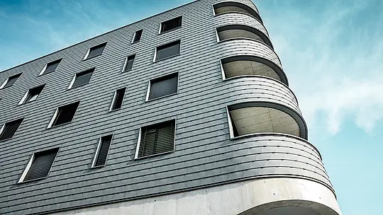 Détail de la Bernerhaus à Buchs, avec son angle arrondi remarquable et ses balcons aux lignes fluides. La façade, composée de bardeaux PREFA gris souris P.10, épouse la structure du bâtiment à la forme nette comme une peau d'aluminium finement travaillée. La surface texturée accentue les courbes douces et crée un jeu d'ombre et de lumière saisissant. Grâce à la légèreté, la résistance aux intempéries et la durabilité de l'aluminium PREFA, on obtient une enveloppe de bâtiment homogène et nécessitant peu d'entretien – une prouesse technique et un véritable chef-d'œuvre architectural.