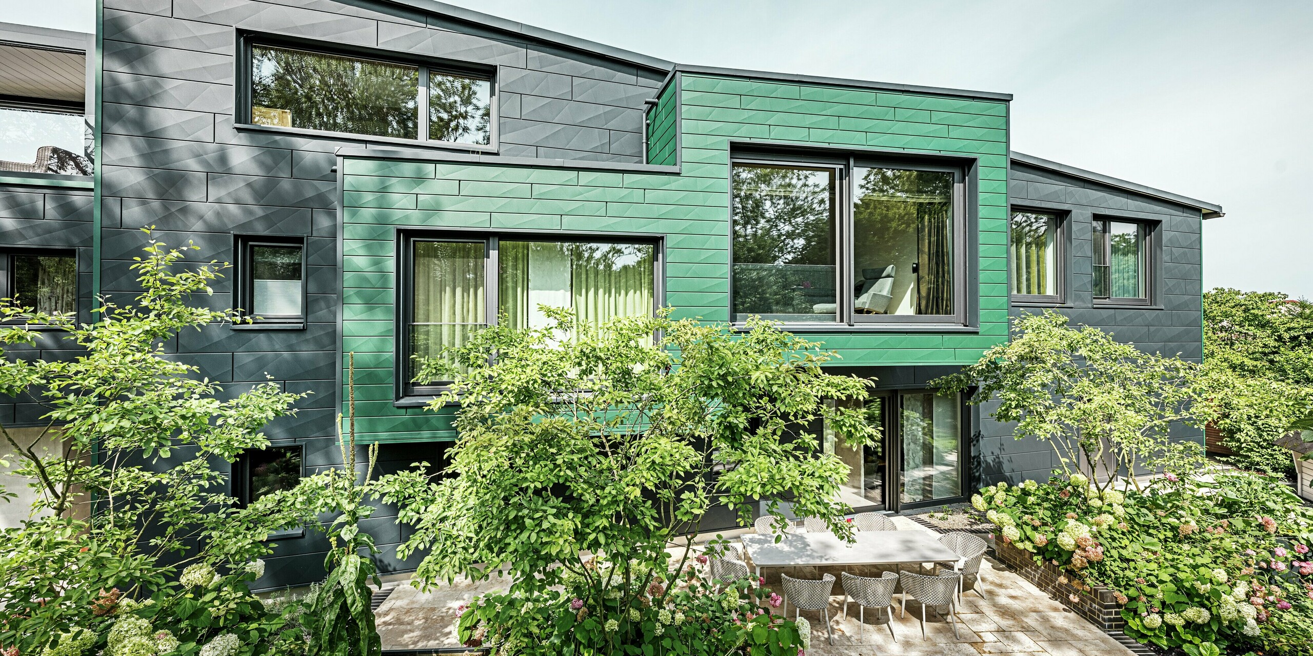 Das Einfamilienhaus in Türkheim besticht durch seine moderne und dynamische Architektur sowie seine markante Fassade aus PREFA Siding X-Paneelen in den Farben Anthrazit und Moosgrün. Die klaren Linien der Paneele verleihen dem Gebäude eine lebendige Struktur. Im Vordergrund befindet sich ein Gartenbereich mit üppigen Hortensien und einer stilvollen Terrasse. Die pflegeleichte und wetterbeständige Aluminiumfassade schützt das Gebäude und verleiht ihm eine zeitlose Eleganz.