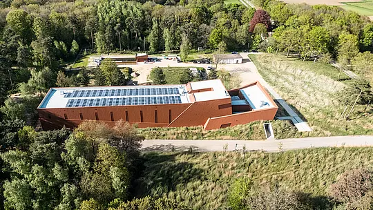 Vue aérienne de la station de pompage d'eau potable WML à Lottum, une infrastructure remarquable s'intégrant harmonieusement à un paysage verdoyant de bois et de prairies. Le bâtiment allongé, avec sa façade distinctive en aluminium PREFA Siding.X à la finition chaleureuse couleur rouille, se dresse comme un repère sculptural. Parmi ses caractéristiques remarquables, on note la structure clairement définie du bâtiment, les modules photovoltaïques intégrés sur le toit et la surface de façade finement facettée. Durables, légers et recyclables, les panneaux en aluminium PREFA offrent une résistance exceptionnelle aux intempéries, une efficacité énergétique durable et une enveloppe de bâtiment nécessitant peu d'entretien.