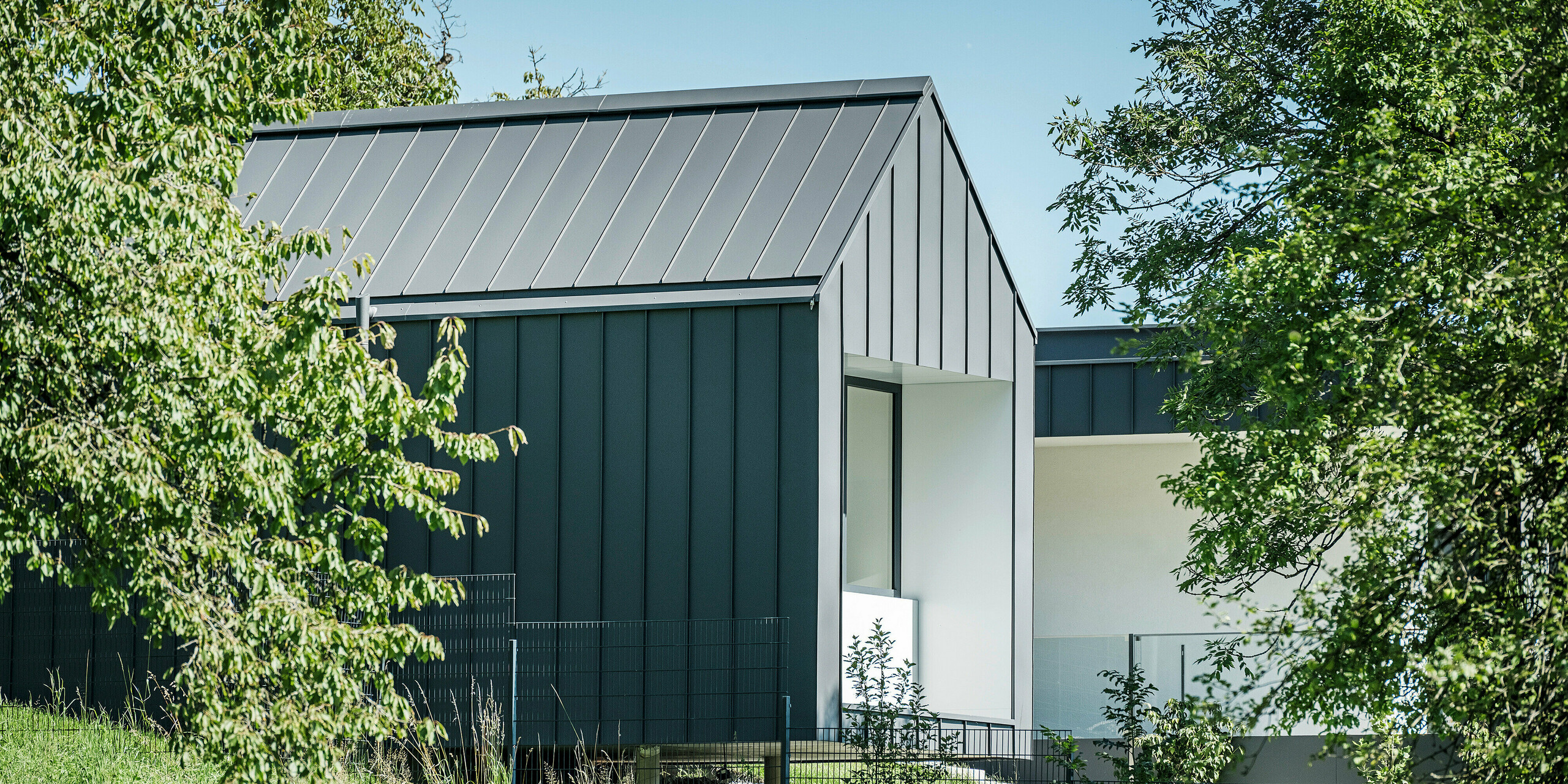Pohled skrz svěží zeleň rodinného domu v rakouském Pogier, vybaveného střechou a fasádou od PREFA v produktu PREFALZ v P.10 tmavě šedá. Jasné linie a tmavé barvy hliníkových prvků tvoří elegantní kontrast se světlou omítnutou fasádou v krytých prostorách a přírodním prostředí. Lodžie nabízí chráněný venkovní prostor, který zdůrazňuje propojení vnitřního a venkovního prostoru a zajišťuje příjemnou atmosféru bydlení.