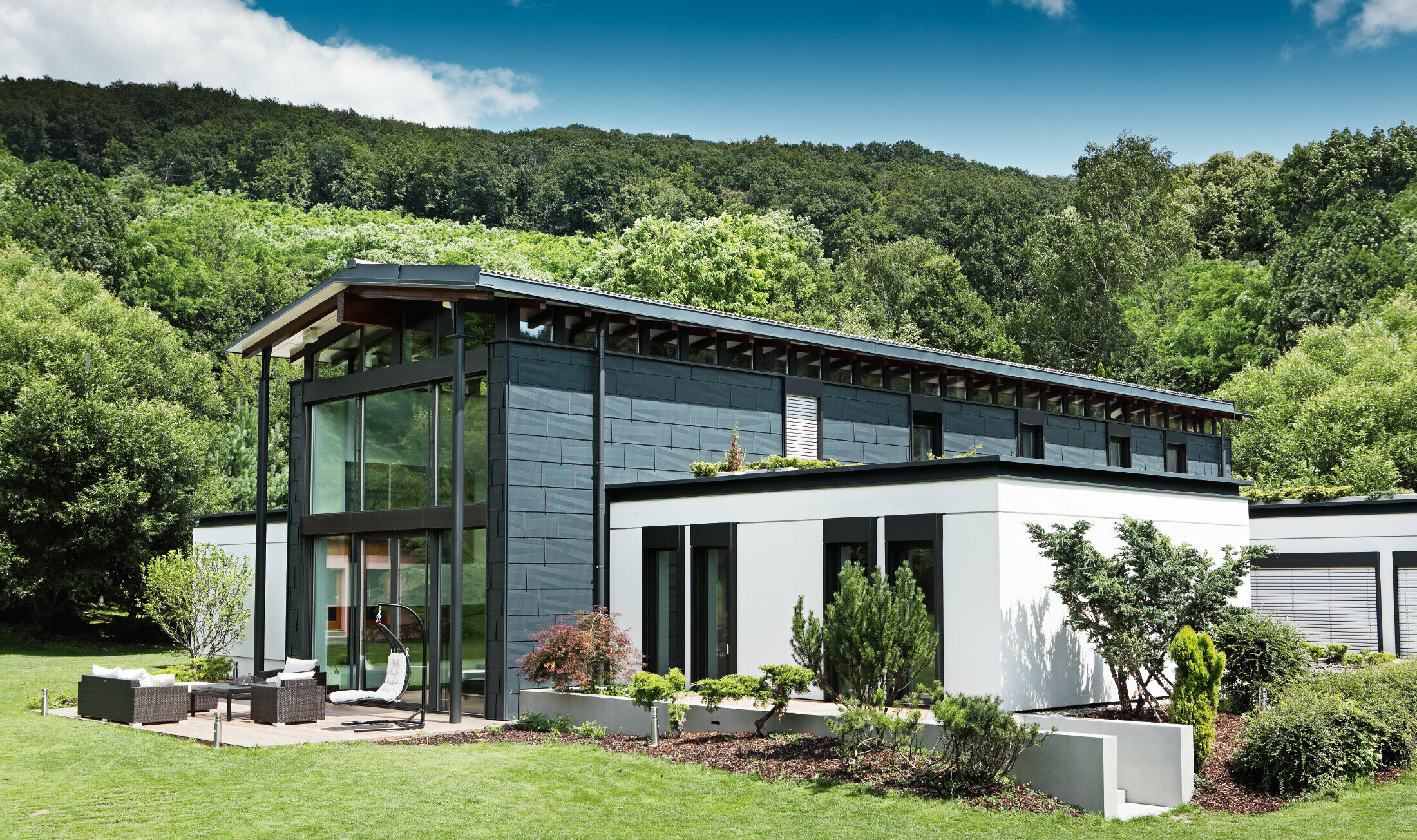 Modernes Einfamilienhaus in Borinka, Slowakei, mit großen Fensterflächen an der einen Hausseite und dem Fassadenpaneel in Anthrazit verkleidet; ein Zubau ist mit einer weißen Putzfassade bekleidet;