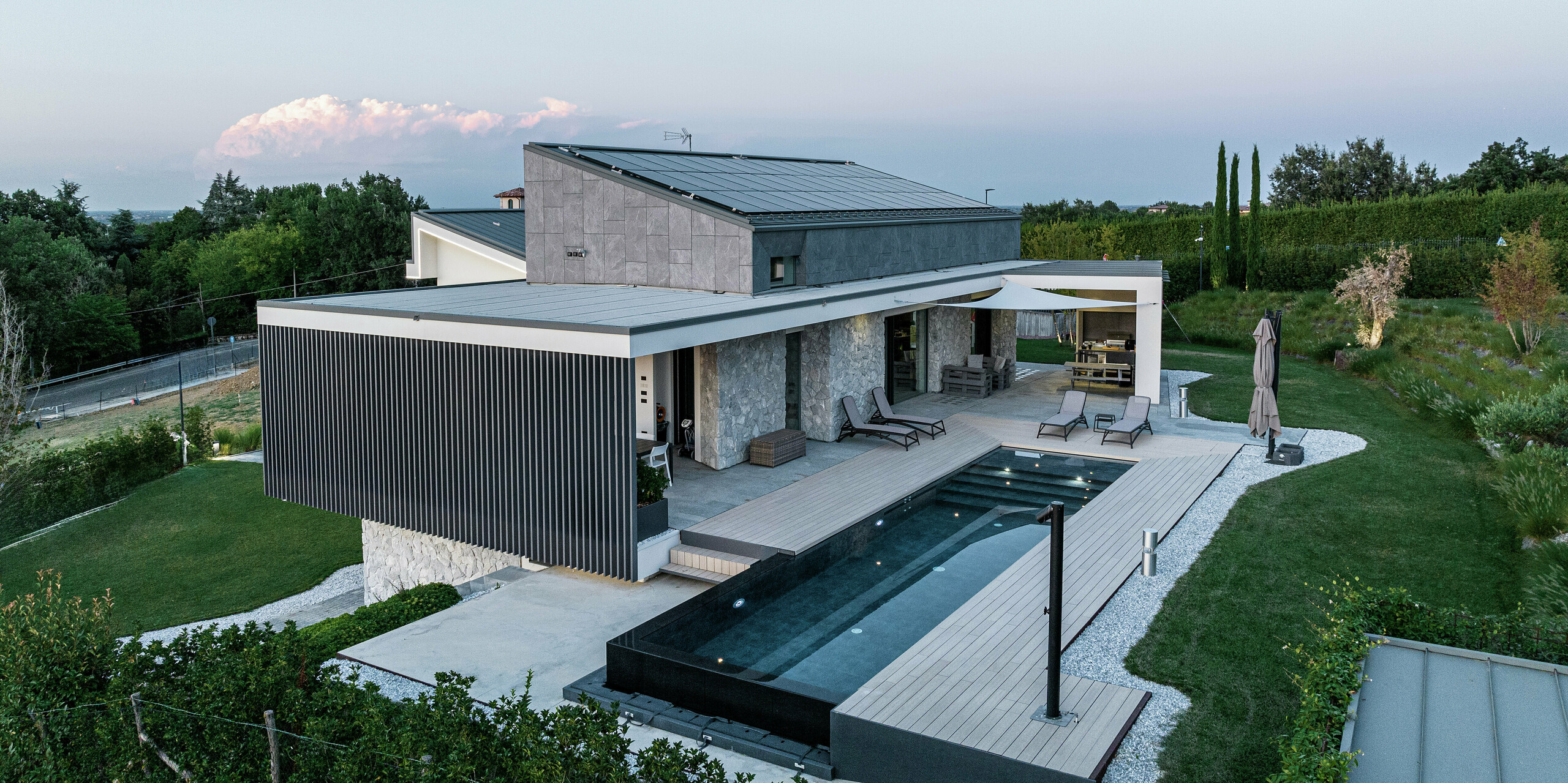 Seitenansicht des luxuriösen Wohnhauses mit Blick auf das elegante PREFALZ Dach in P.10 Anthrazit. Die nachhaltige Aluminiumlösung von PREFA garantiert ein langlebiges, pflegeleichtes Dach in modernem Design. Die großzügige, überdachte Terrasse mit Loungebereich und Infinity-Pool schafft eine exklusive Wohlfühlatmosphäre im Einklang mit der umgebenden Natur. Die Photovoltaikanlage versorgt das Haus mit umweltfreundlichem Strom aus Sonnenenergie.